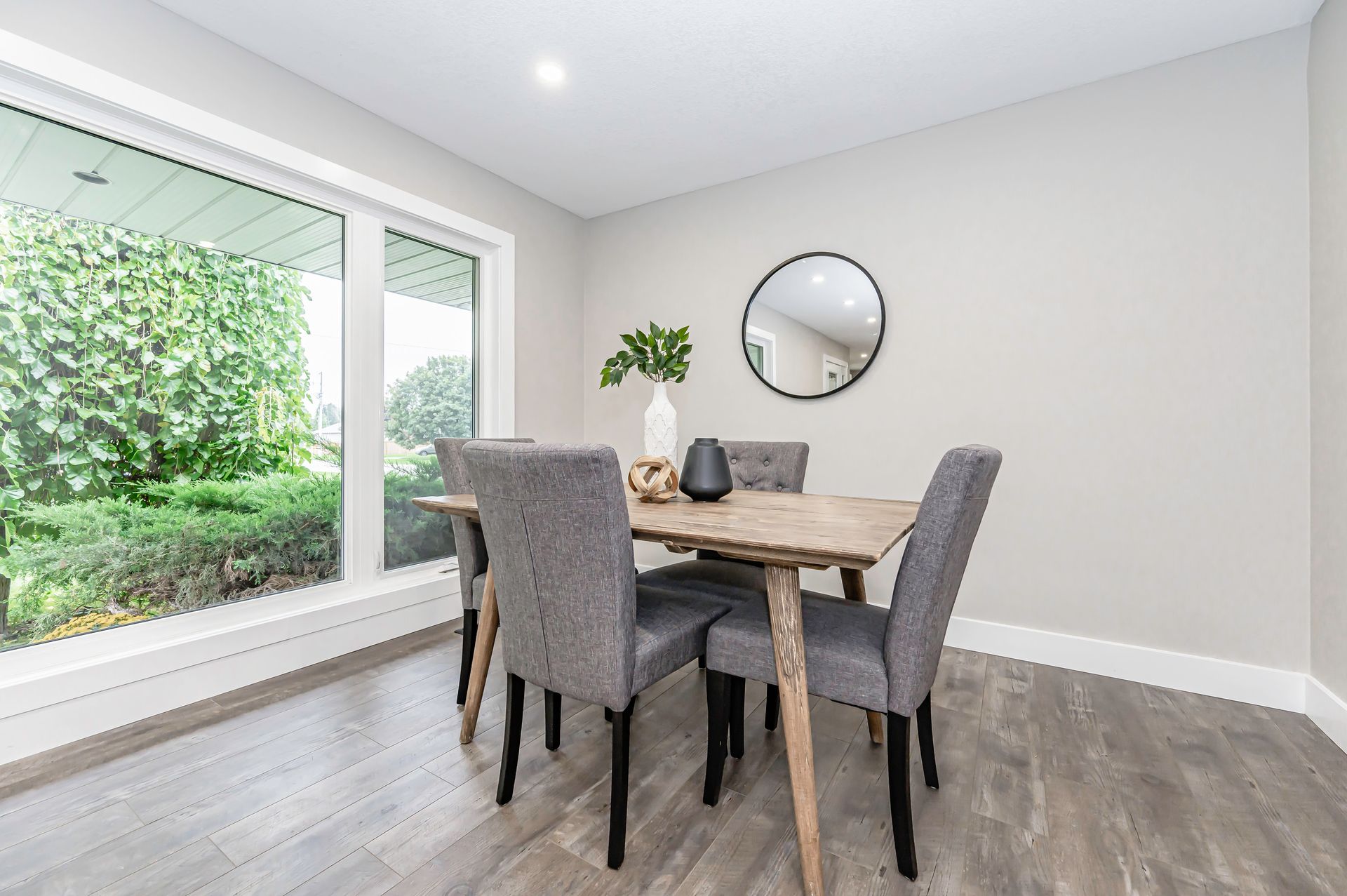 Dining room with wooden table, gray chairs, large window, and round mirror.