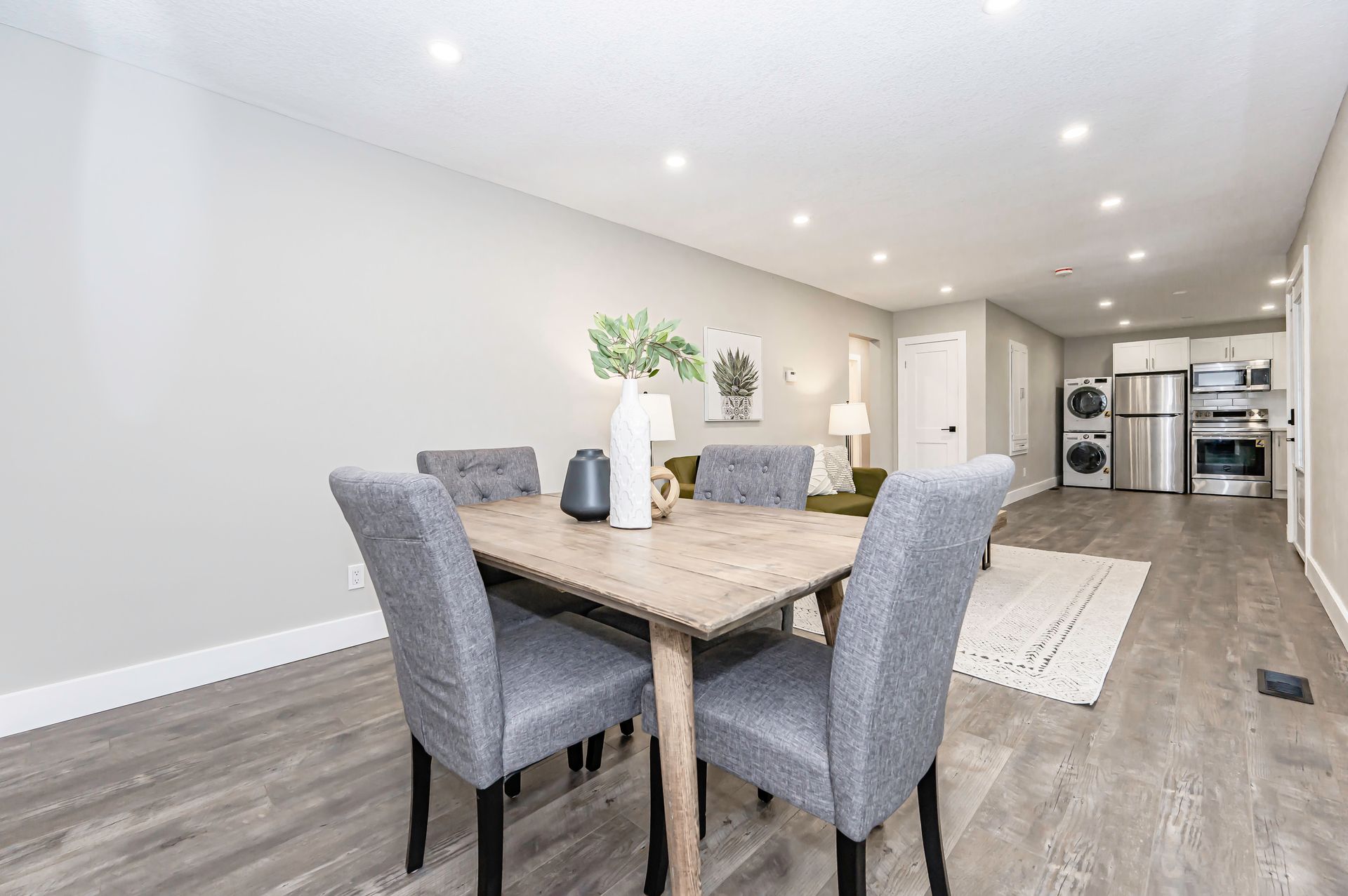 Dining area with a table, four gray chairs, and decorative vase. Living area in background.