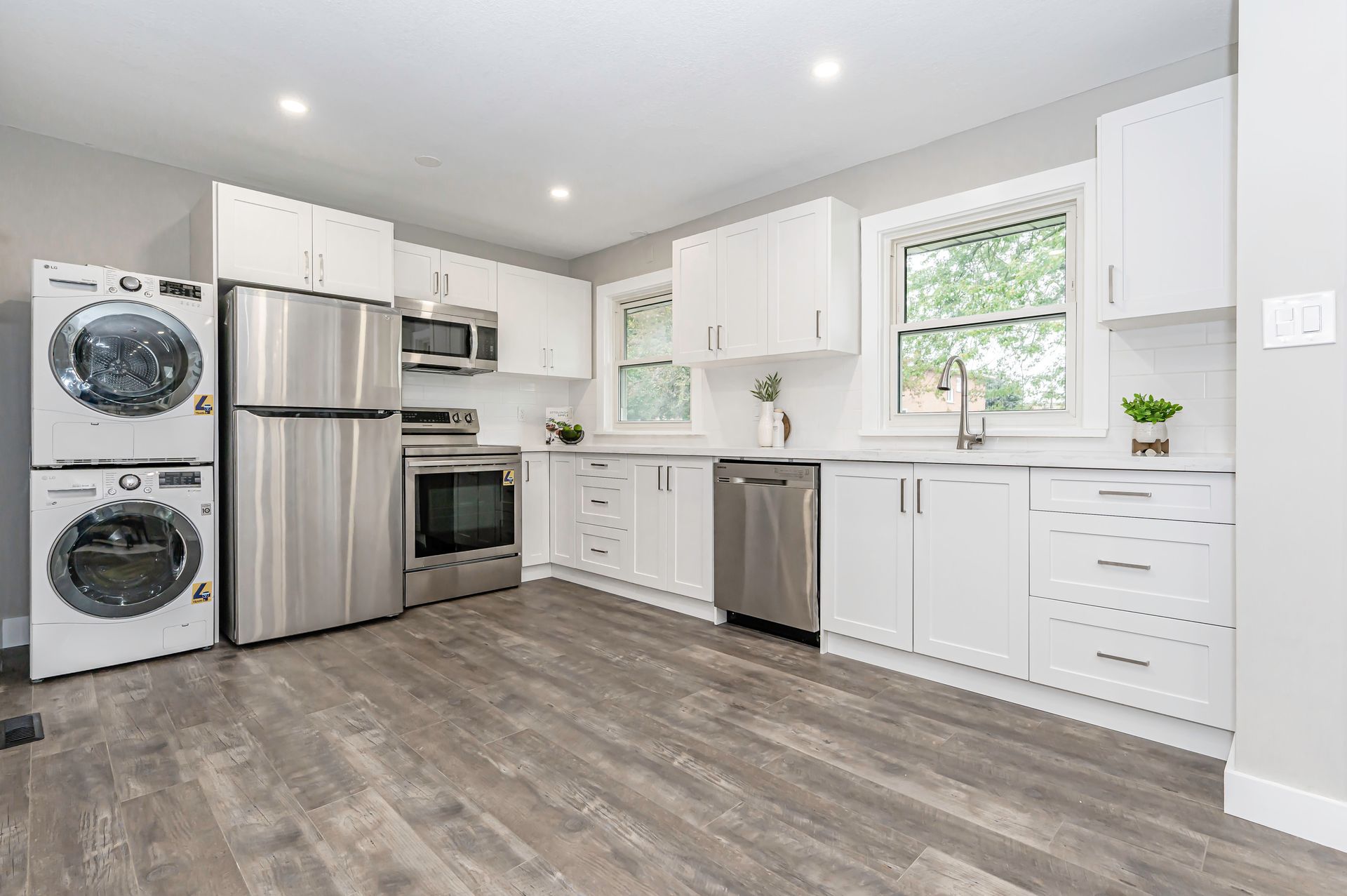 Modern kitchen with white cabinets, stainless steel appliances, and stacked washer/dryer on a gray wood floor.