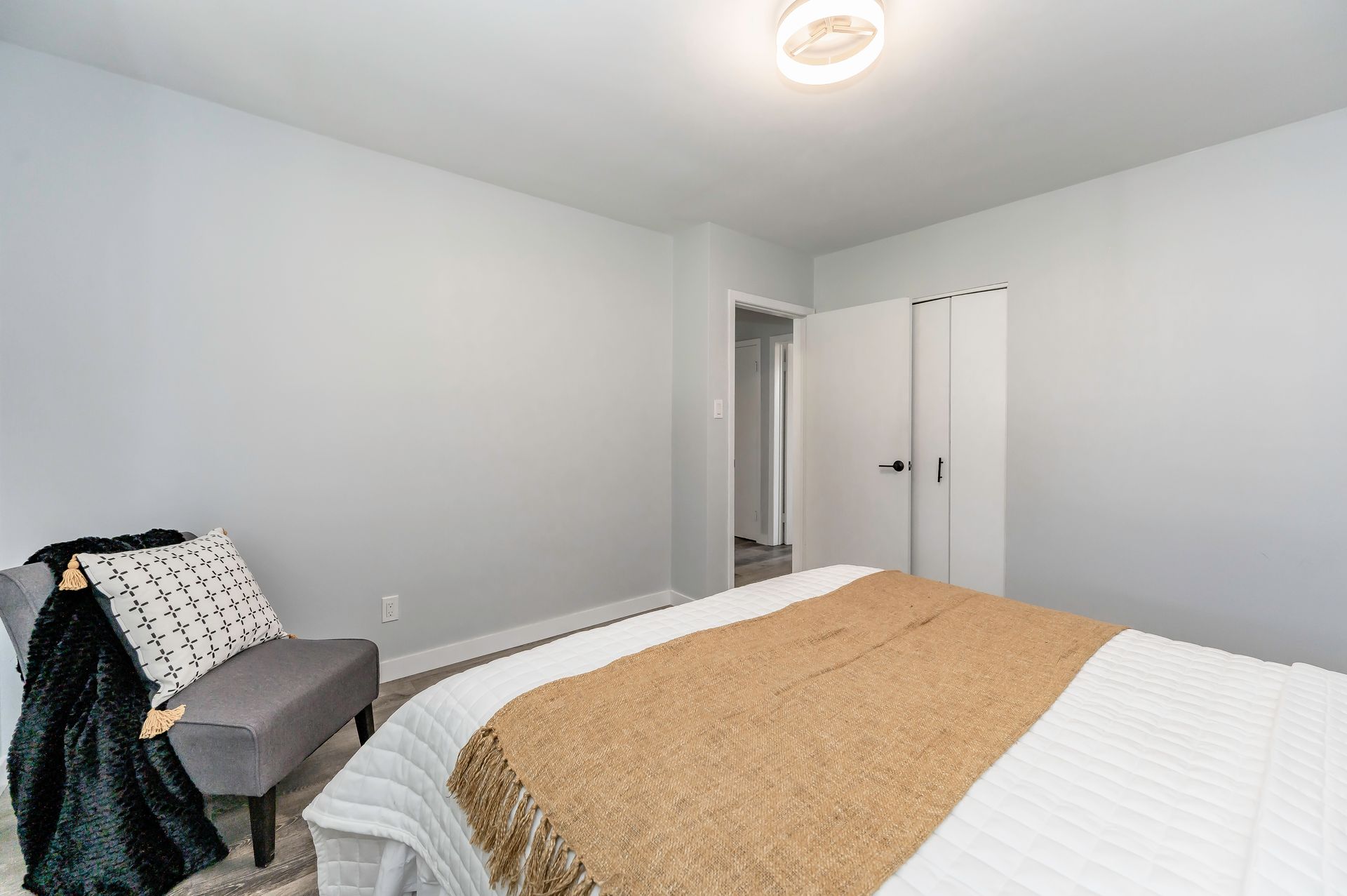 Bedroom with gray walls, bed with gold blanket, gray chair with throw, closet, and ceiling light.
