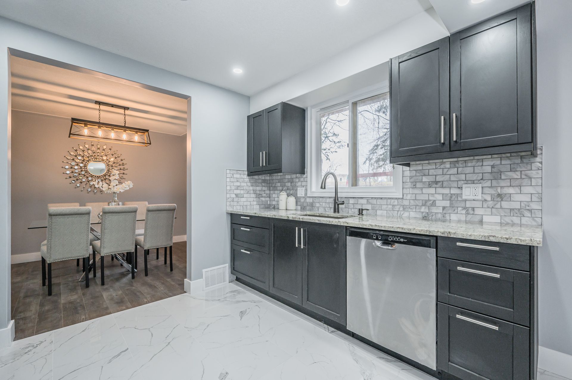 Modern kitchen with dark gray cabinets, stainless steel appliances, and a marble backsplash; a dining room is visible through an opening.