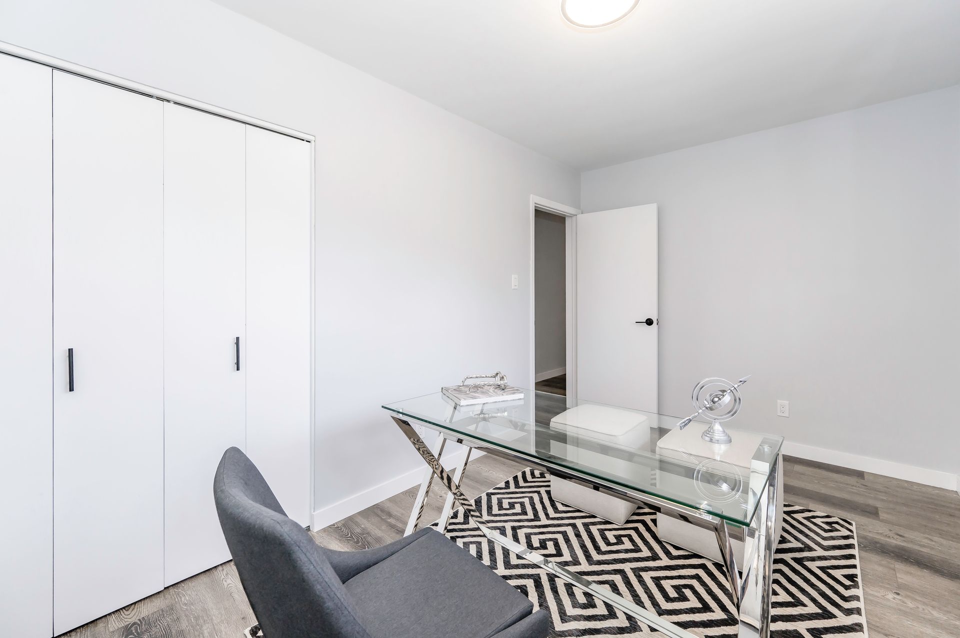 Home office with a glass desk, grey chair, and white walls. A patterned rug and closed closet are also present.