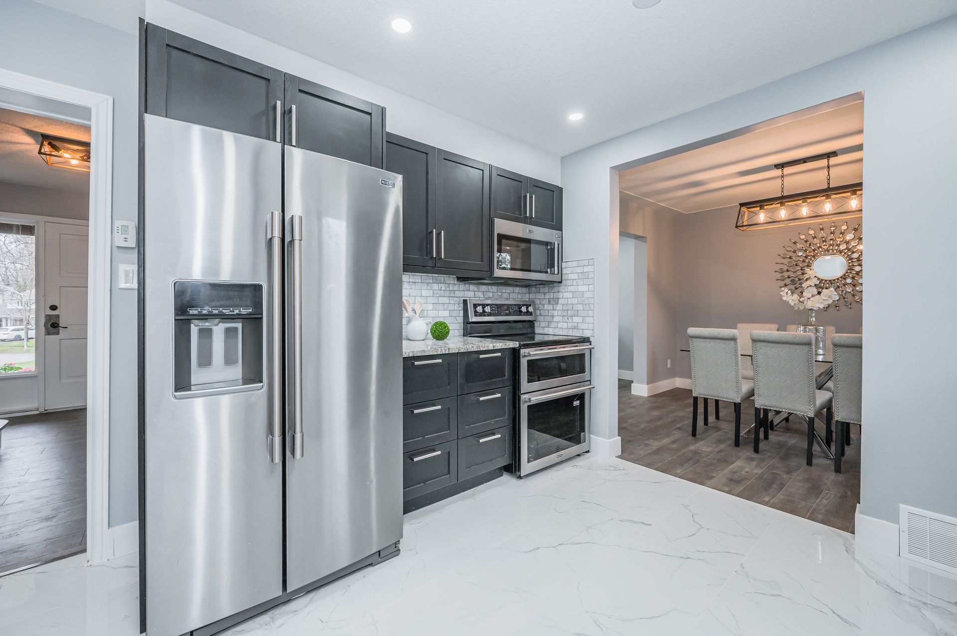 Modern kitchen with stainless steel appliances, dark cabinets, and white tile floor leading to a dining area.