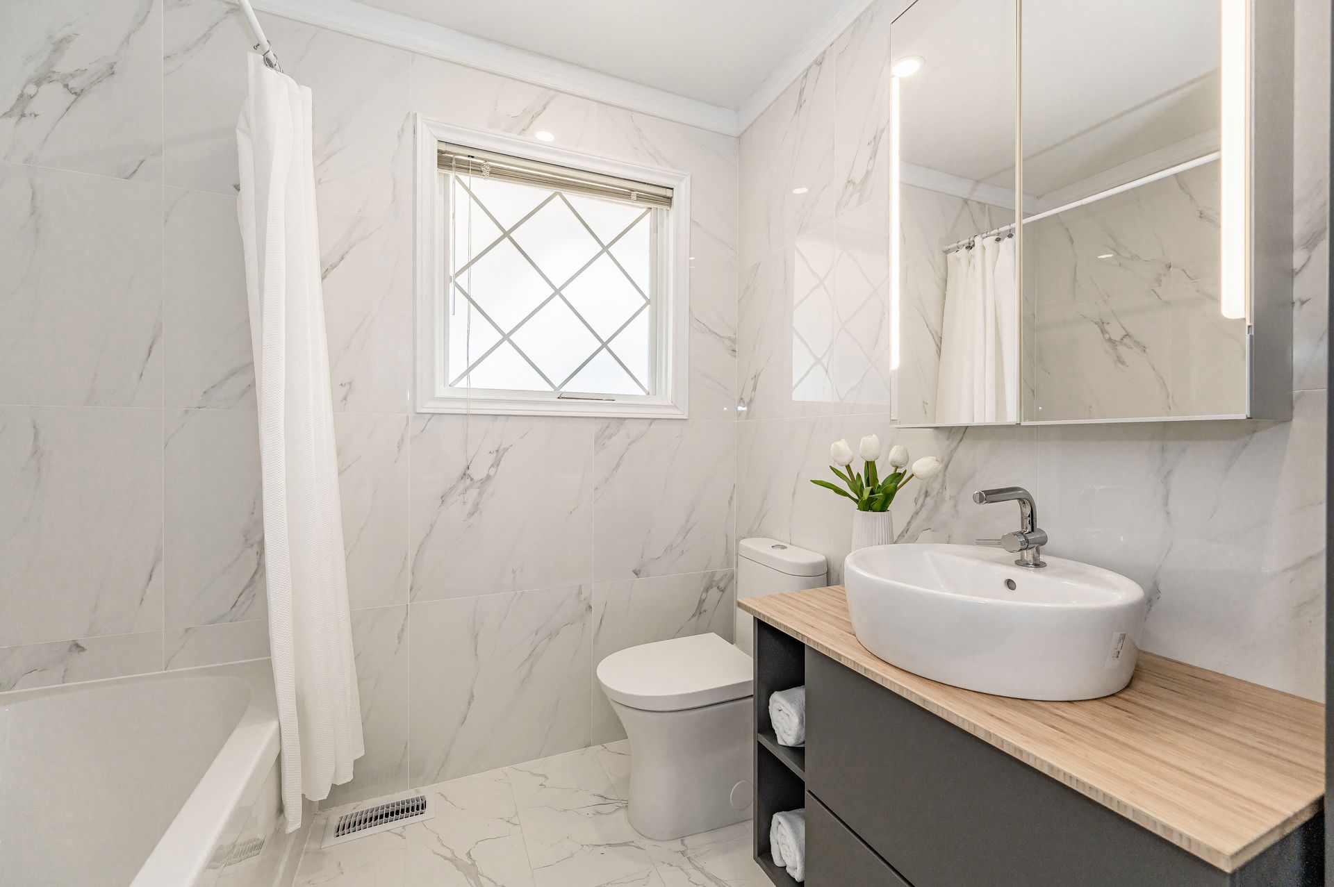 Bathroom with marble-look tile walls, white fixtures, dark gray vanity, and a window with a diamond-patterned pane.