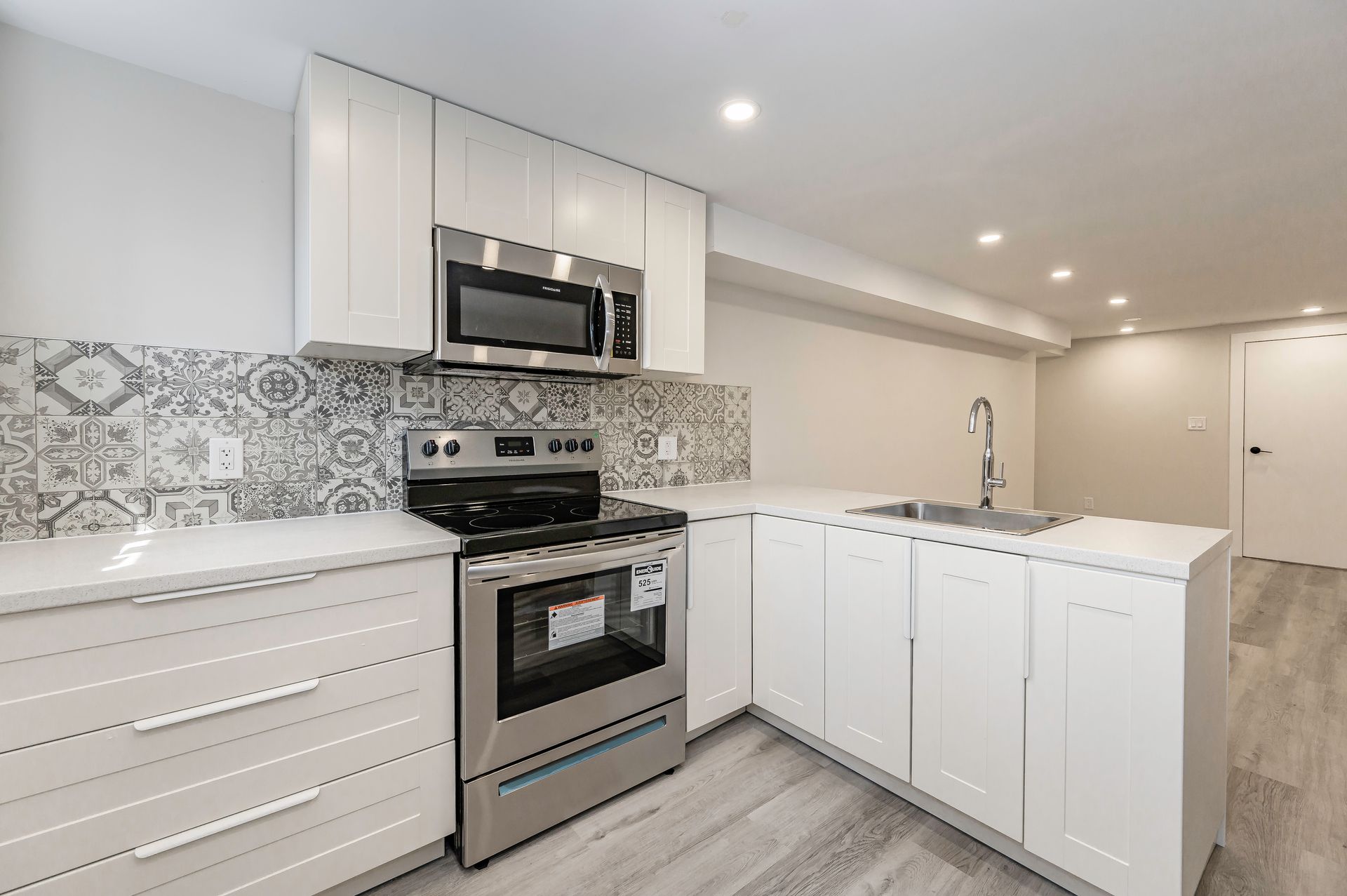 Modern white kitchen with stainless steel appliances, patterned backsplash, and light gray flooring.