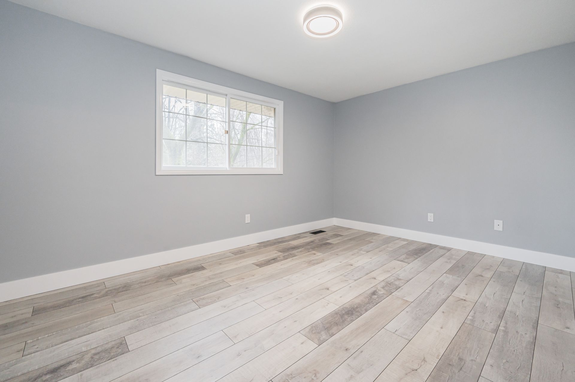 Empty room with grey walls, white trim, and light wood-look flooring. A window and light fixture are visible.