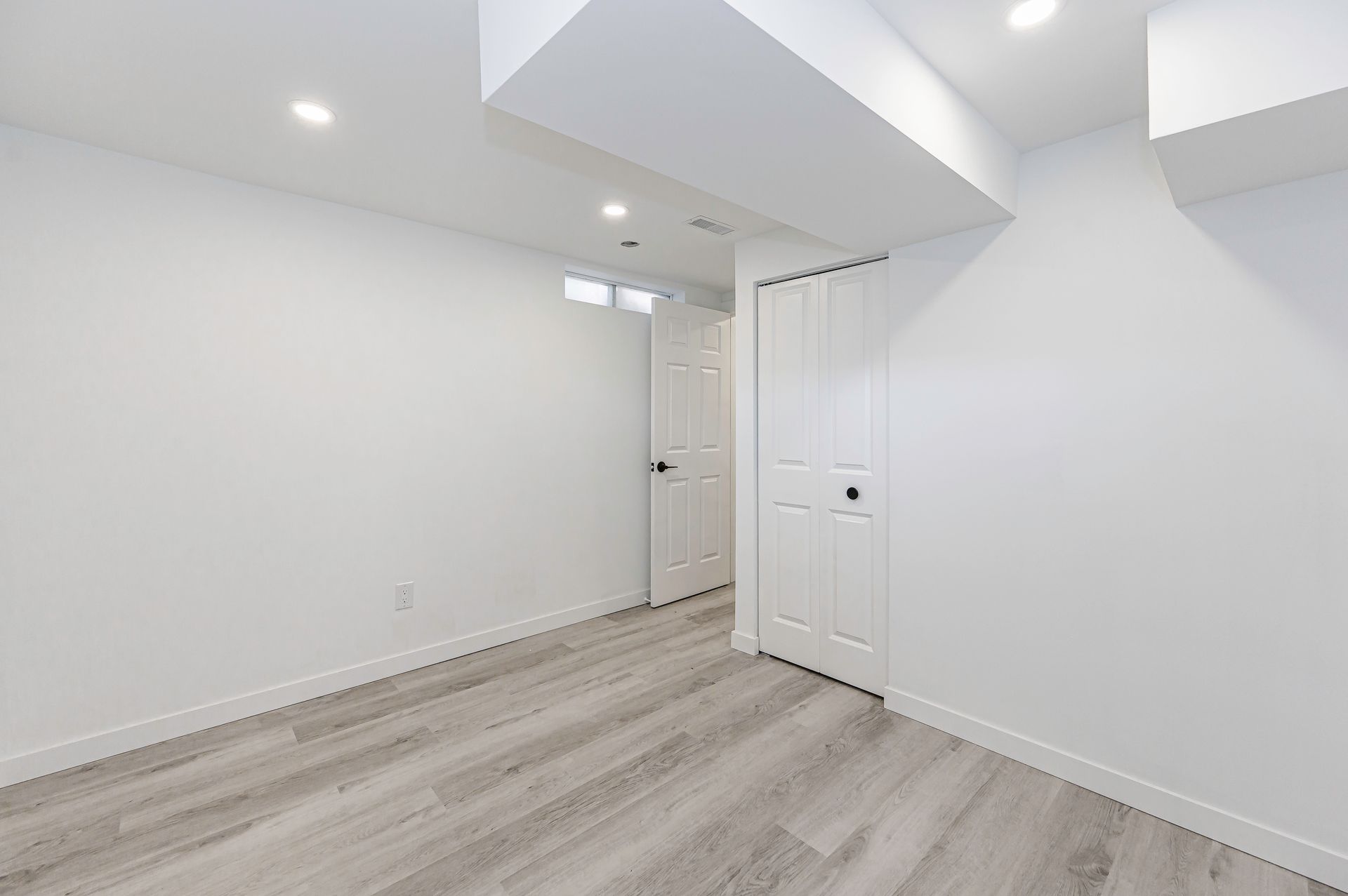 Empty white room with light wood-look flooring, two white doors, and recessed lighting.