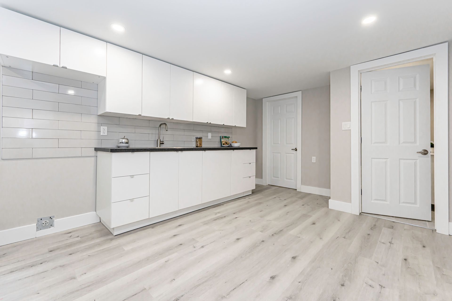 White kitchen cabinets and countertops in a room with light gray walls and flooring, two white doors.