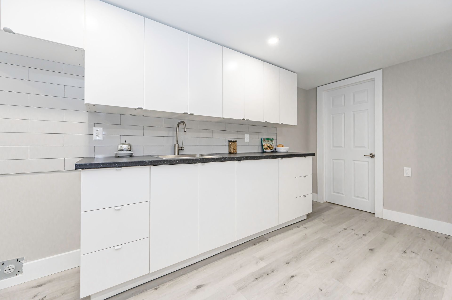 White kitchen cabinets, gray countertop, stainless steel sink, white door, light wood floor.