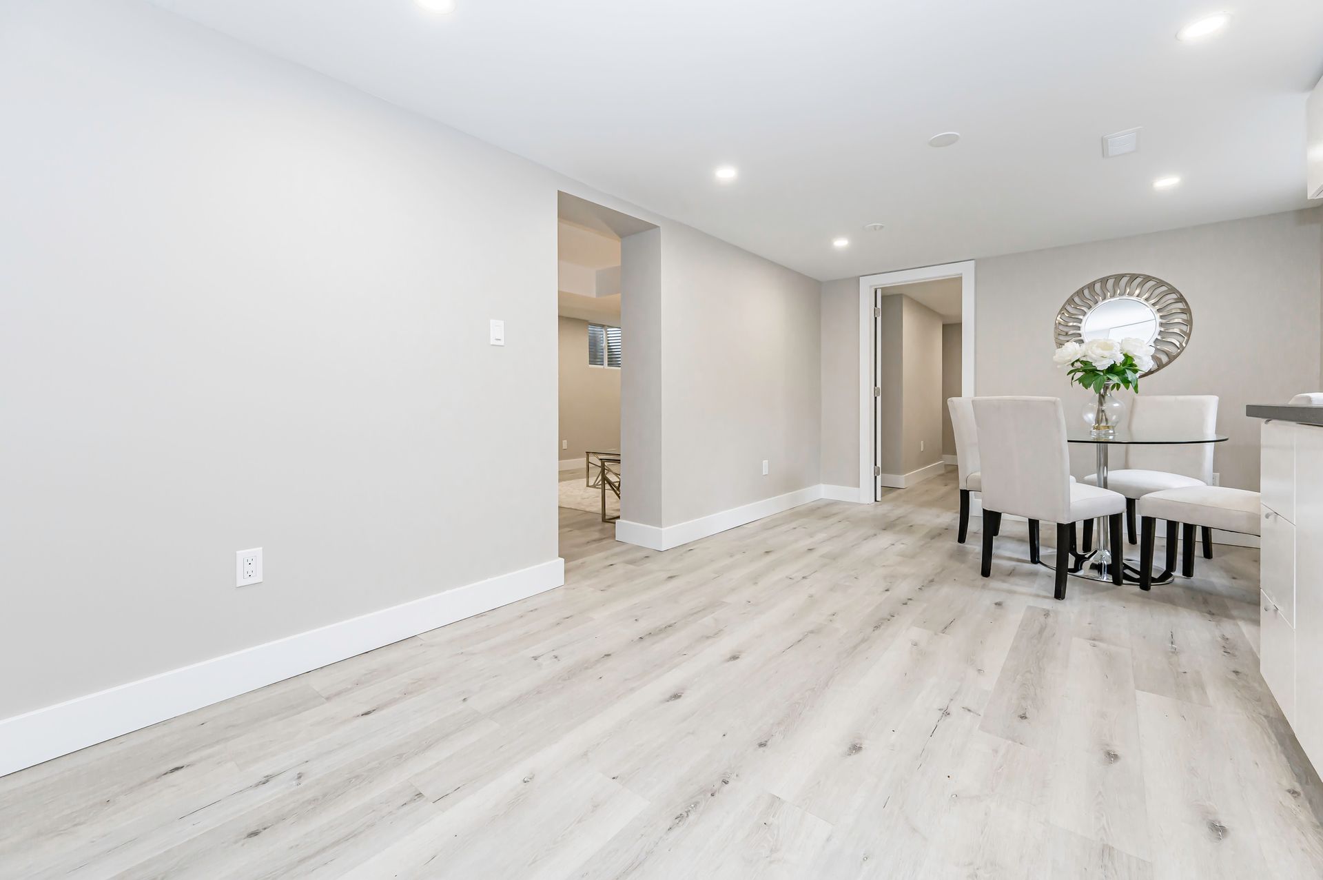 Bright dining area with light wood floors, gray walls, and white trim. A glass table with four chairs sits in the corner.