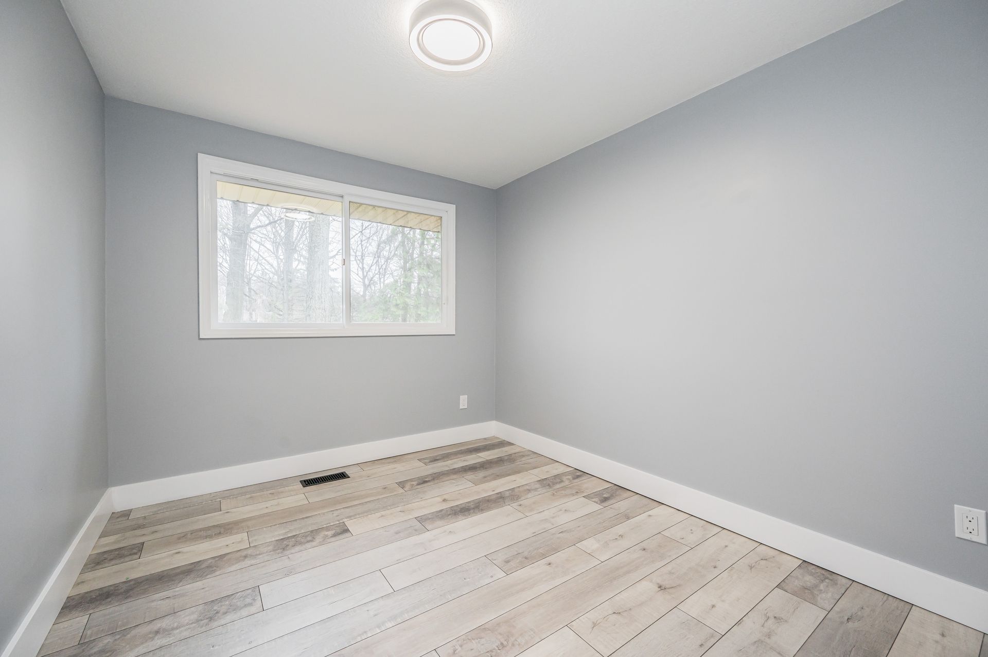 Empty room with light gray walls, white trim, and wood-look flooring, with a window and ceiling light.