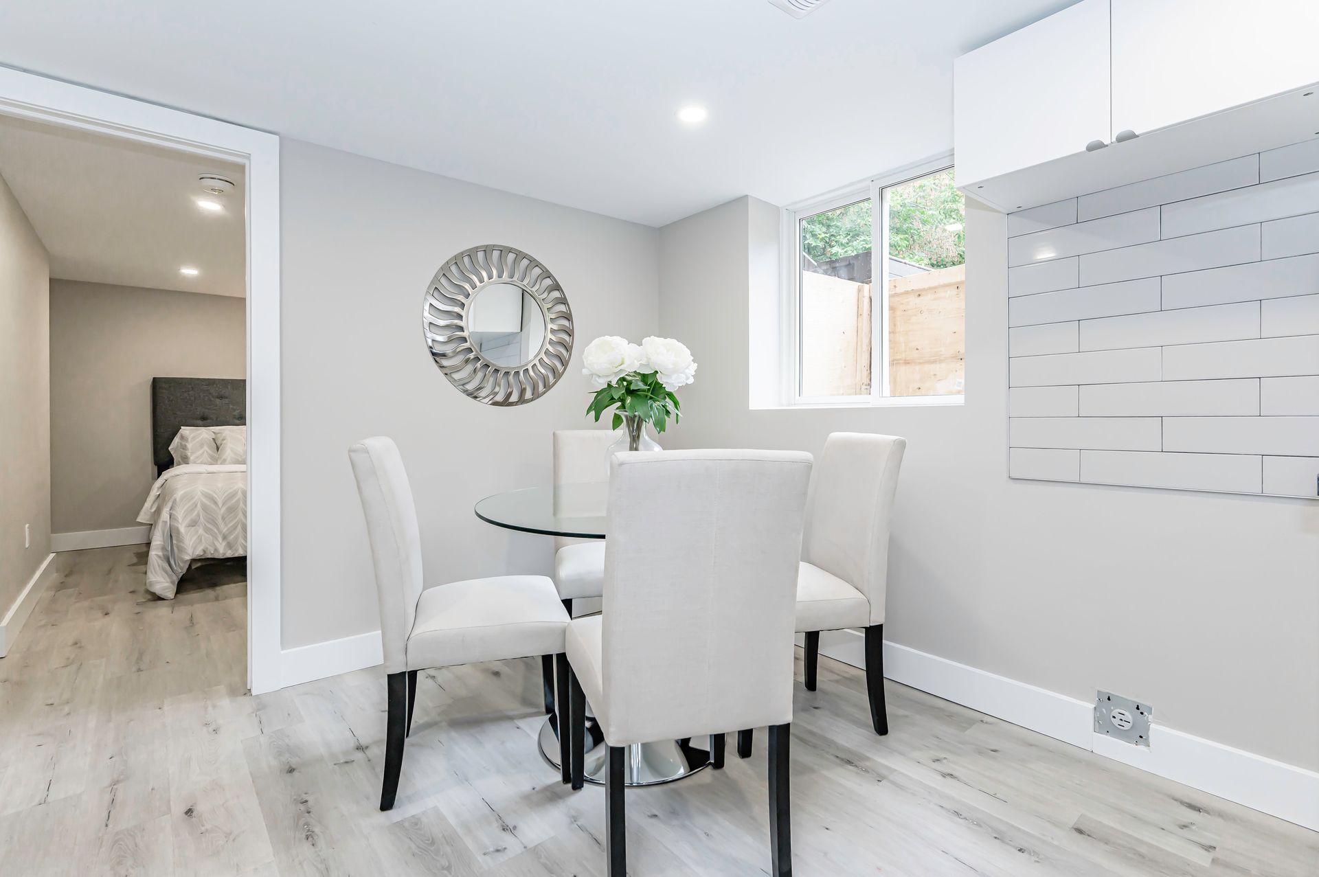 Dining area with a round glass table, four white chairs, and a decorative mirror on the wall.