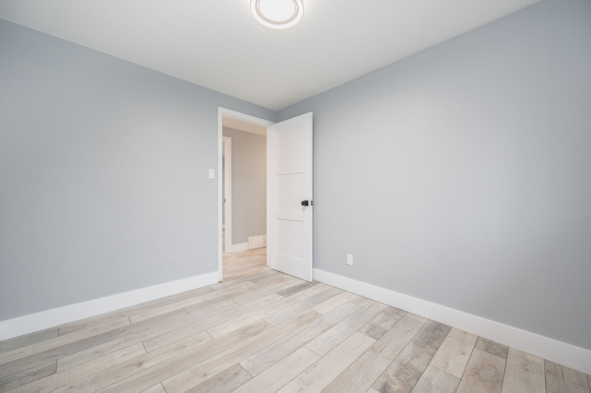 Empty bedroom with light blue walls, white trim, and wood-look flooring; an open door leads to another room.