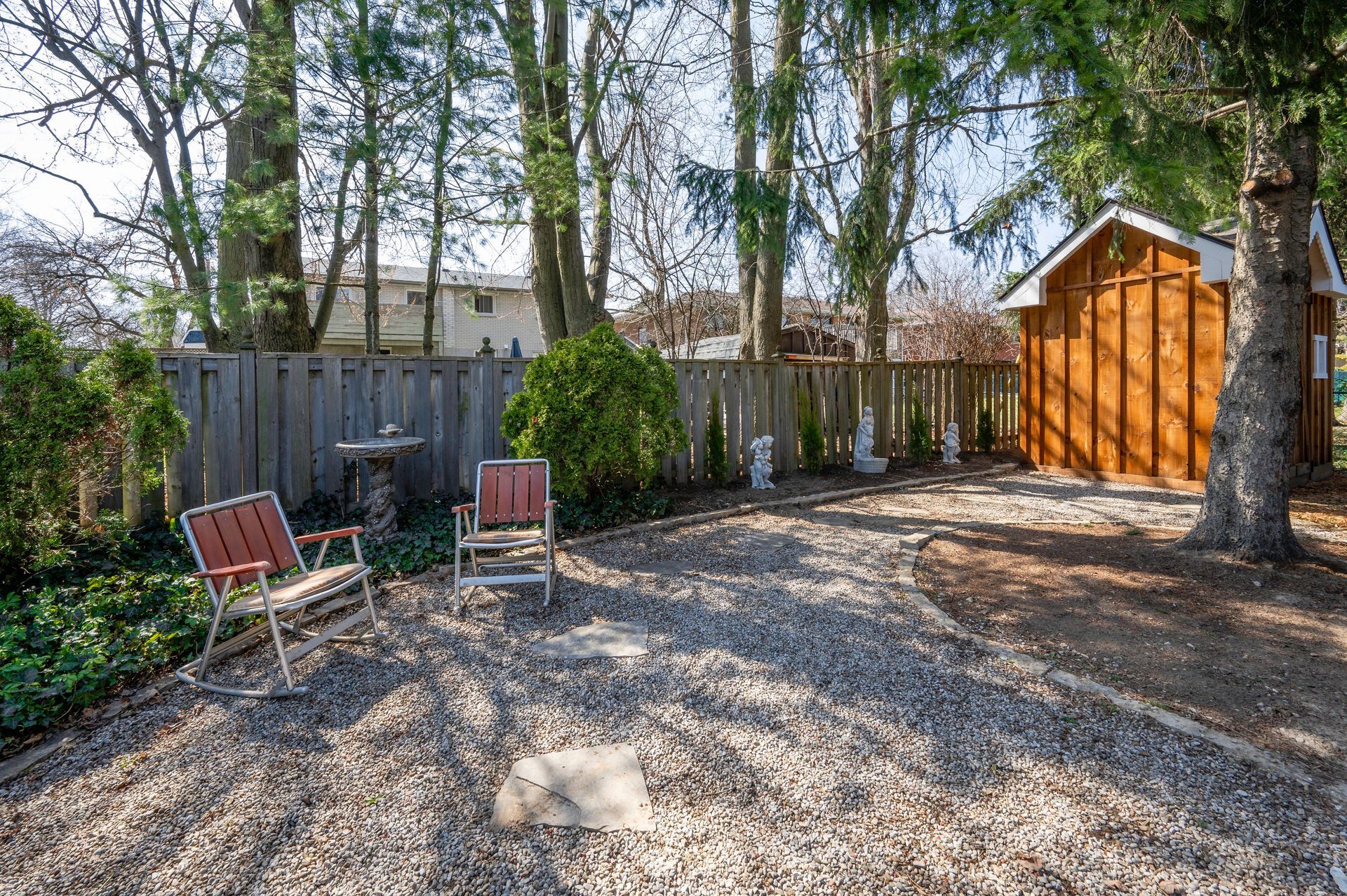 Gravel backyard with two chairs, a shed, and a wooden fence. Trees provide shade on a sunny day.