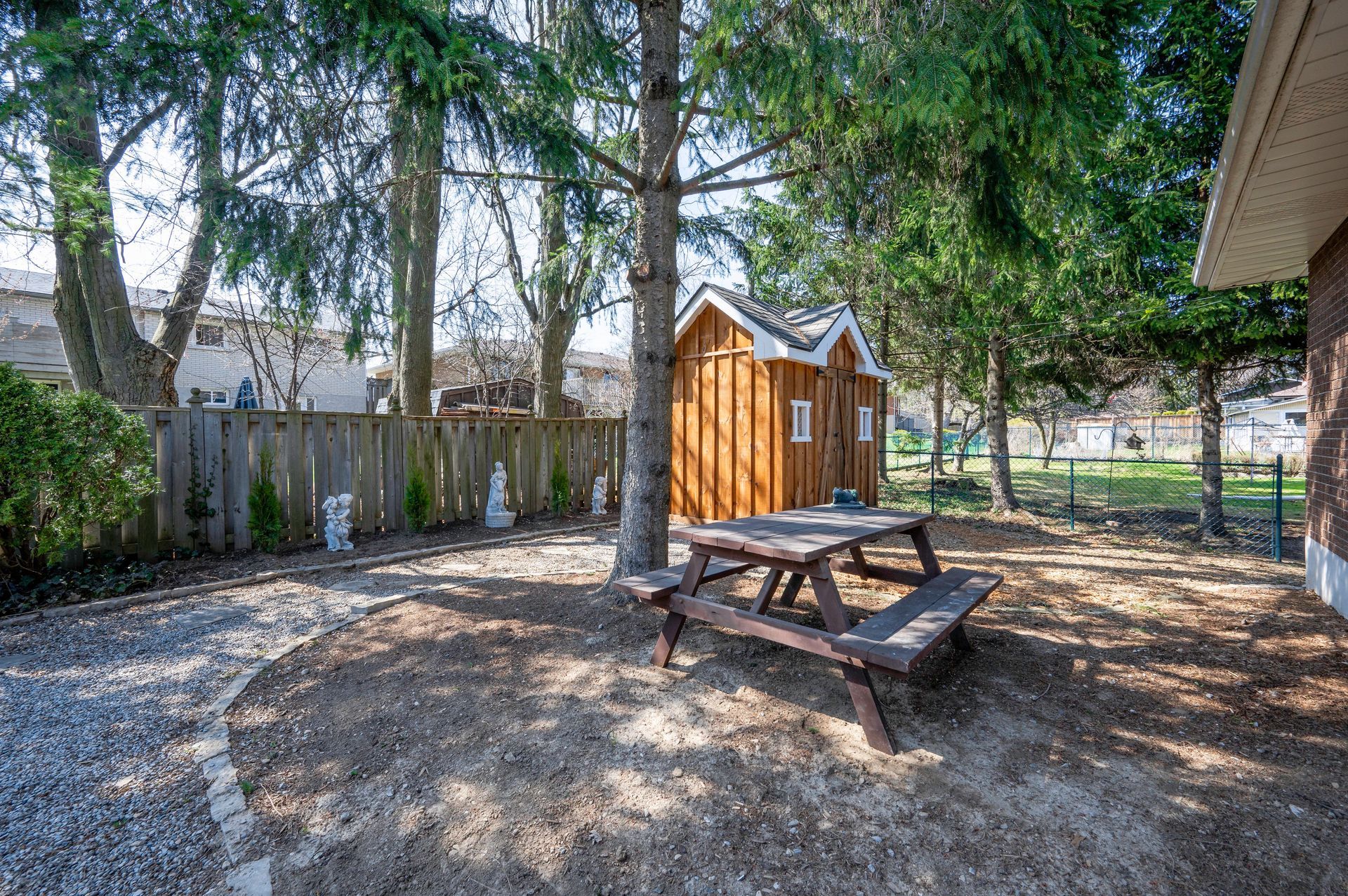 Backyard with picnic table, wooden shed, and trees. Brown picnic table on a dirt path, wooden shed in the background.