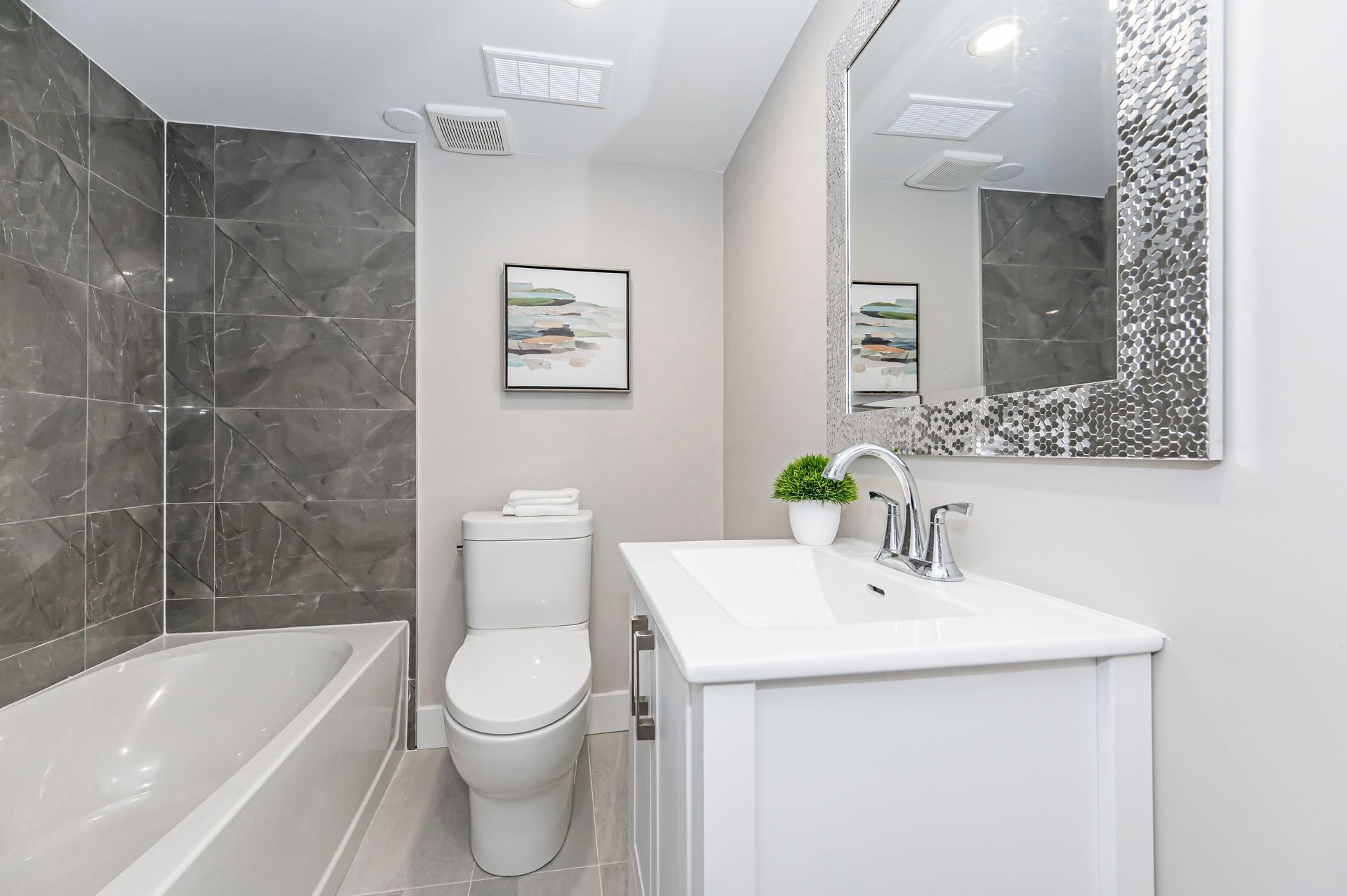 Modern bathroom with white vanity, toilet, and bathtub. Gray tiled shower wall and mirror frame.