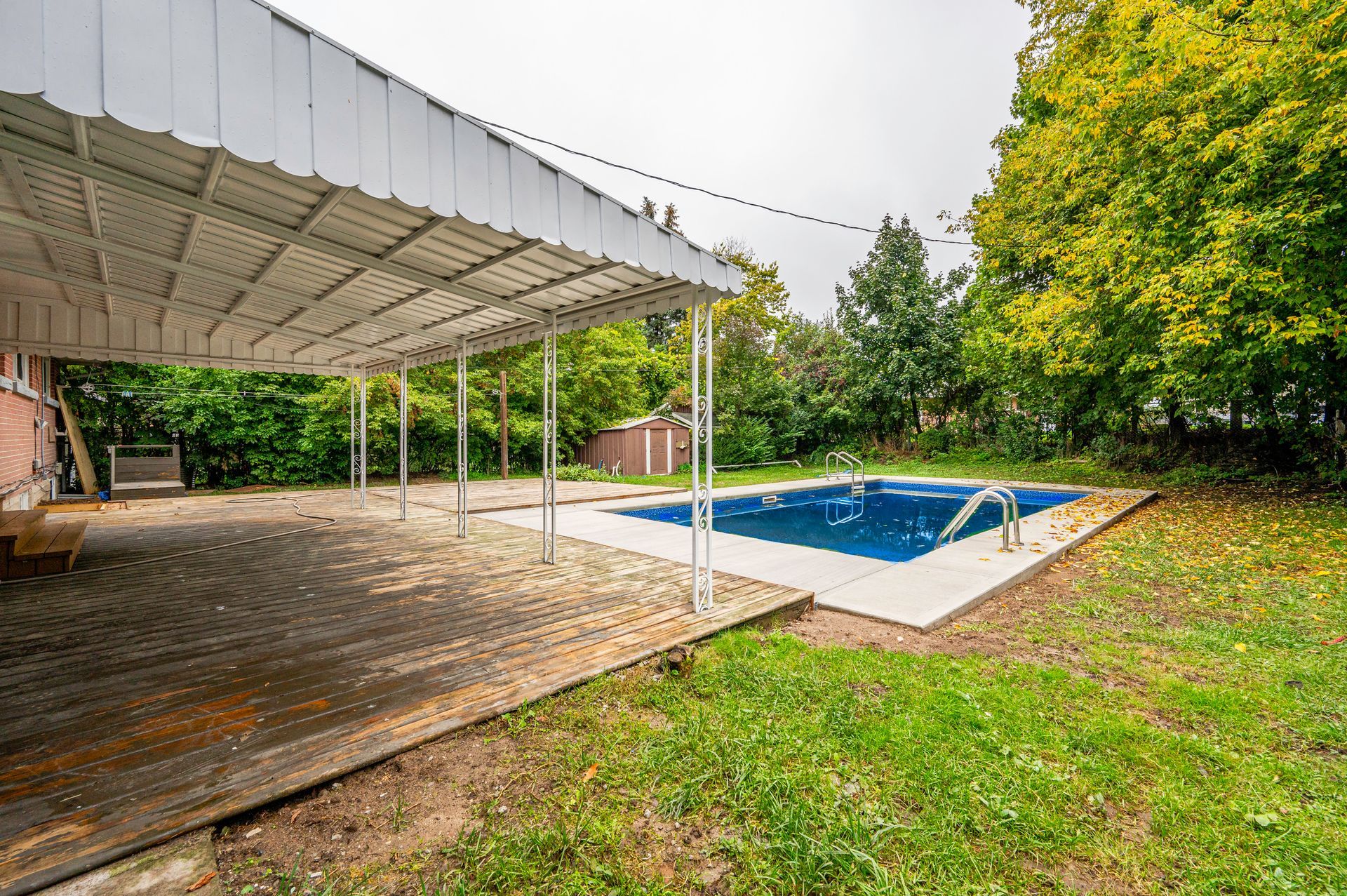 Patio with a pool covered in blue, surrounded by green grass and trees. A white awning covers the patio.