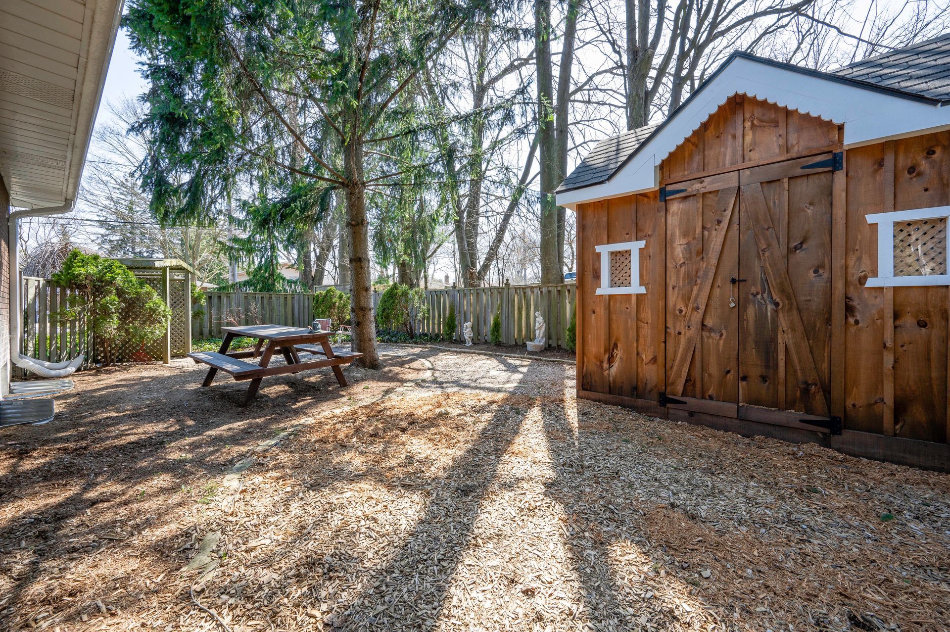 Backyard with wooden shed, picnic table, and gravel ground. Trees and fence in the background.