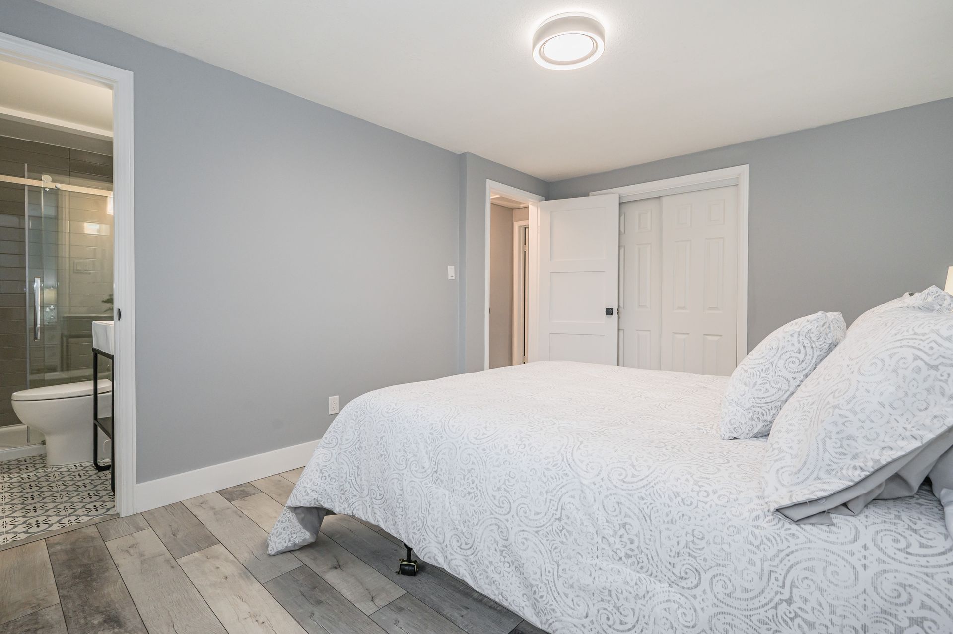 Bedroom with a bed, two white doors, a bathroom, and gray walls and flooring.