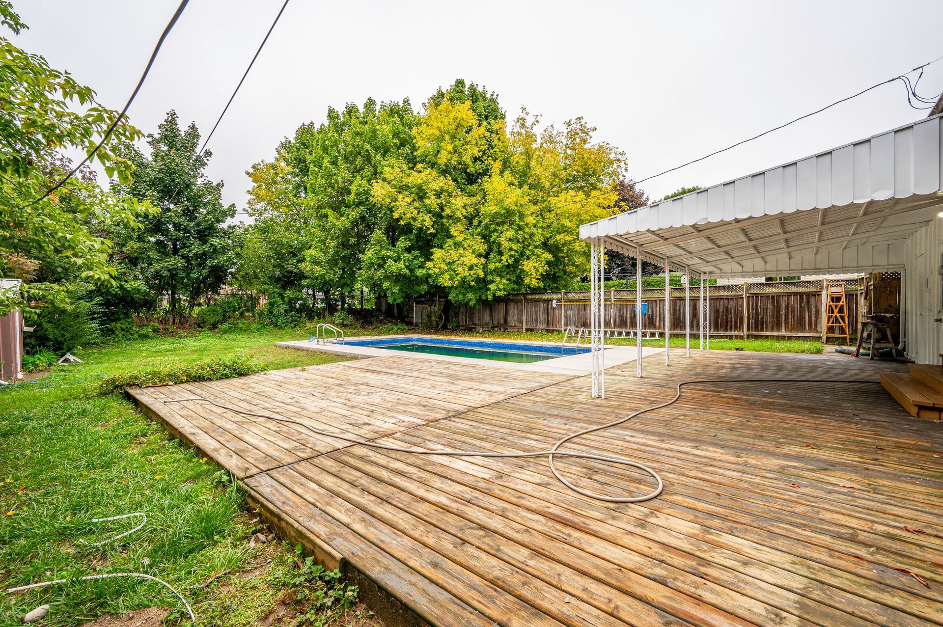 Backyard with a wooden deck, pool, and covered patio under overcast skies.