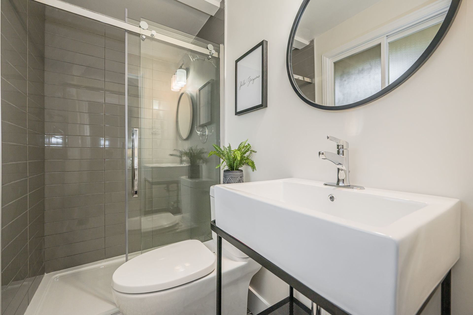 Modern bathroom with a glass shower, white sink, and toilet. Gray tile walls and light gray floor.