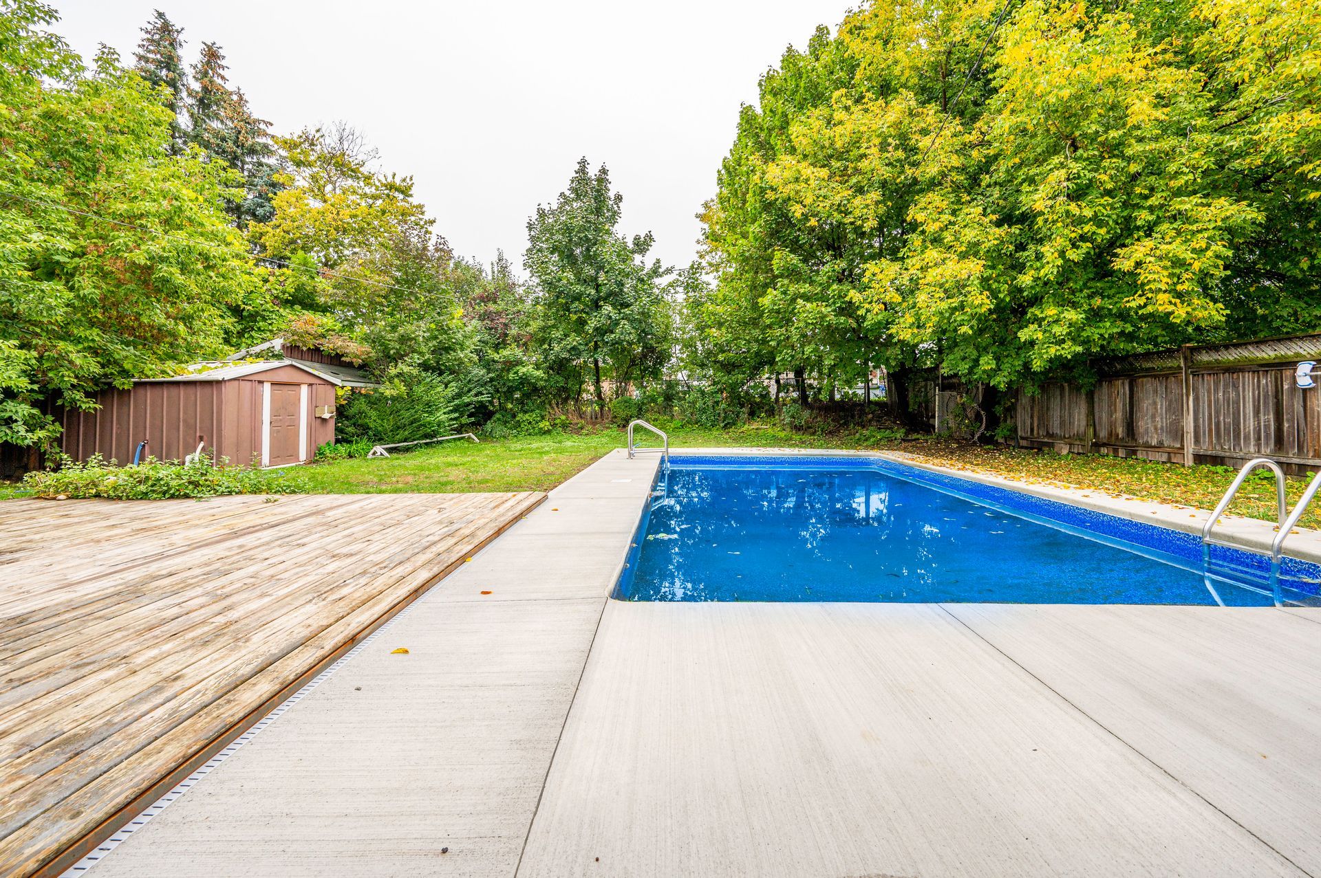 A backyard with a rectangular pool, wooden deck, shed, and trees. Blue pool water.