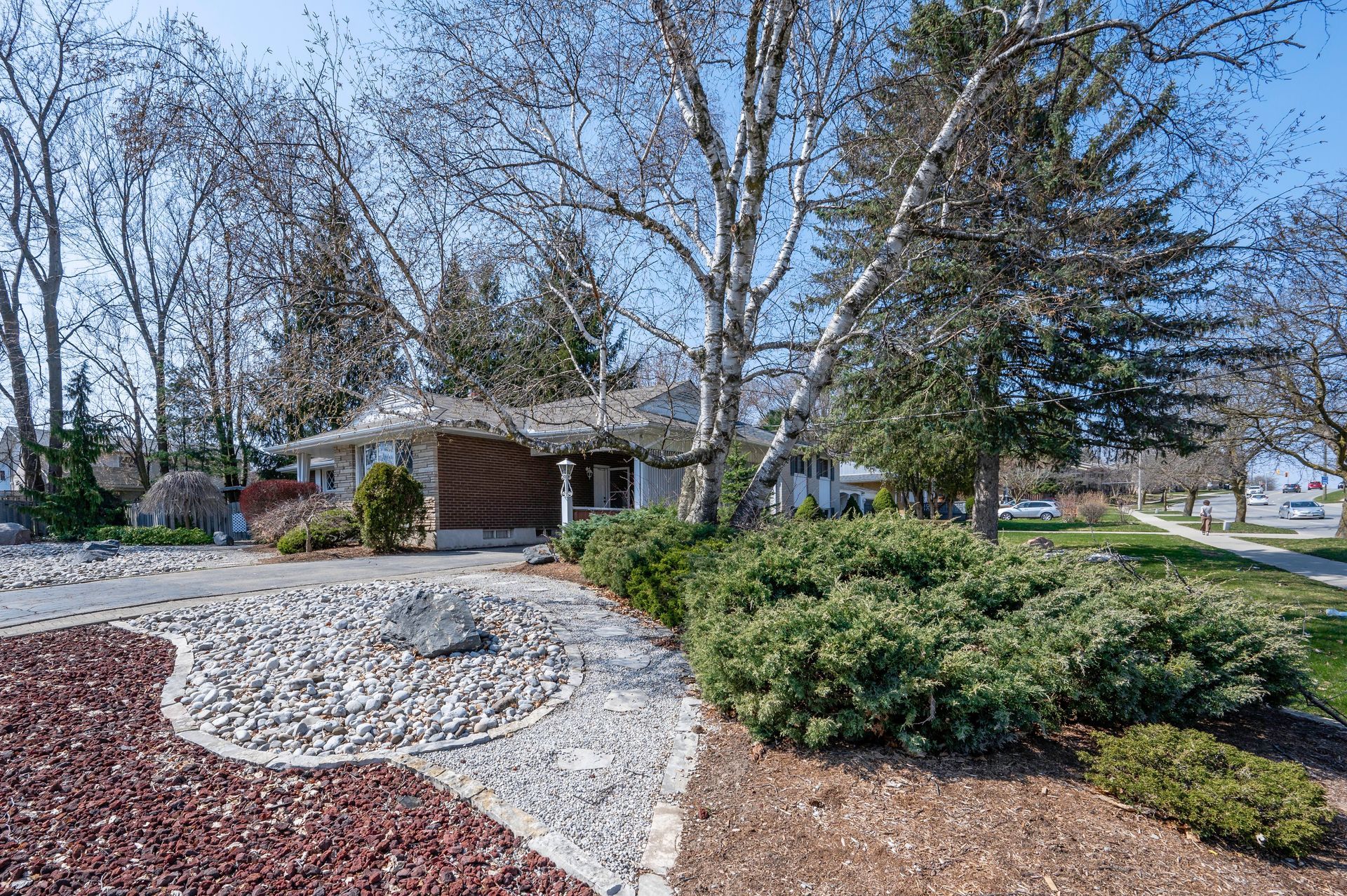 House with circular driveway, stone and bush landscaping. Trees and blue sky overhead.