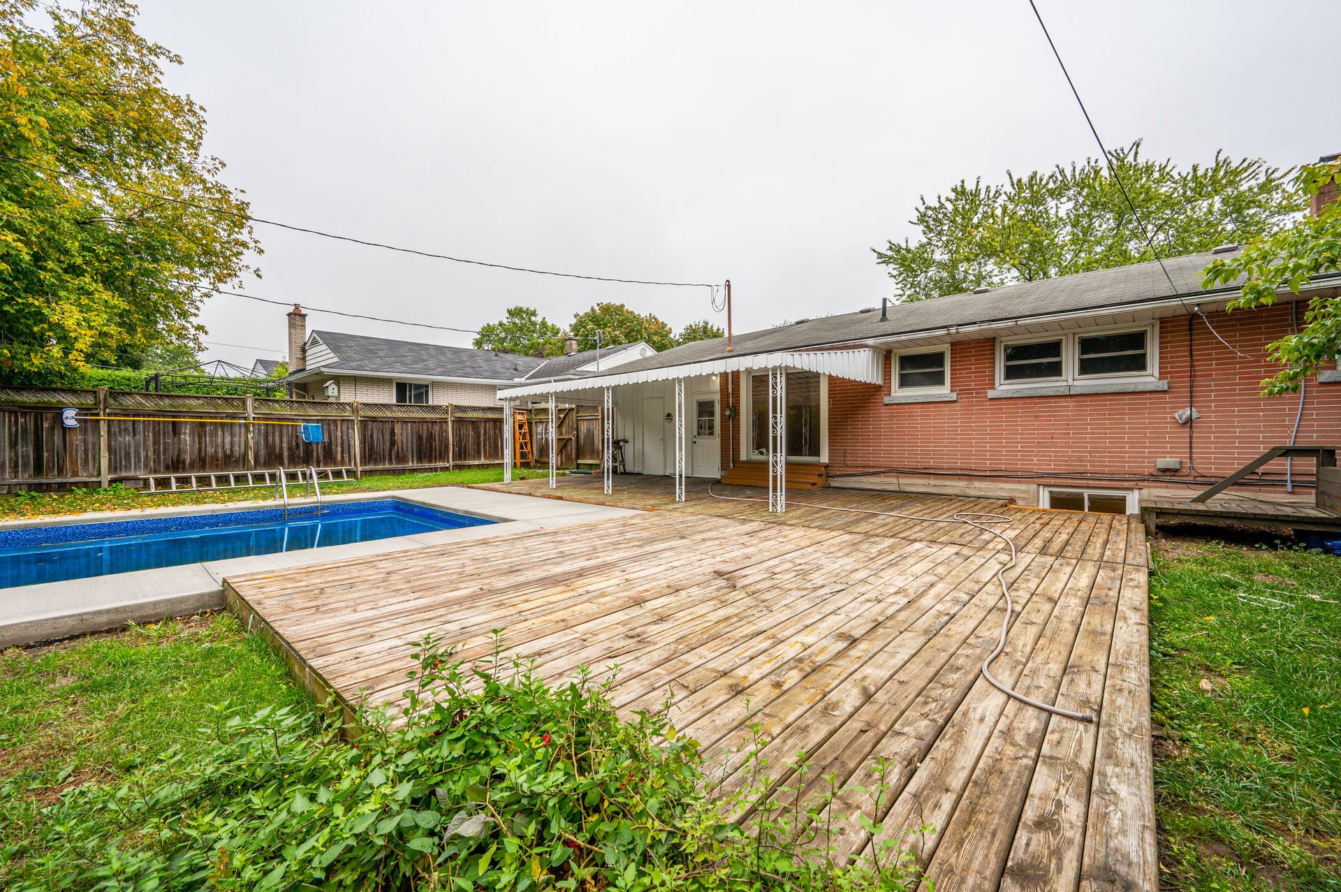 Backyard with wooden deck, pool, and red-brick house under overcast sky.