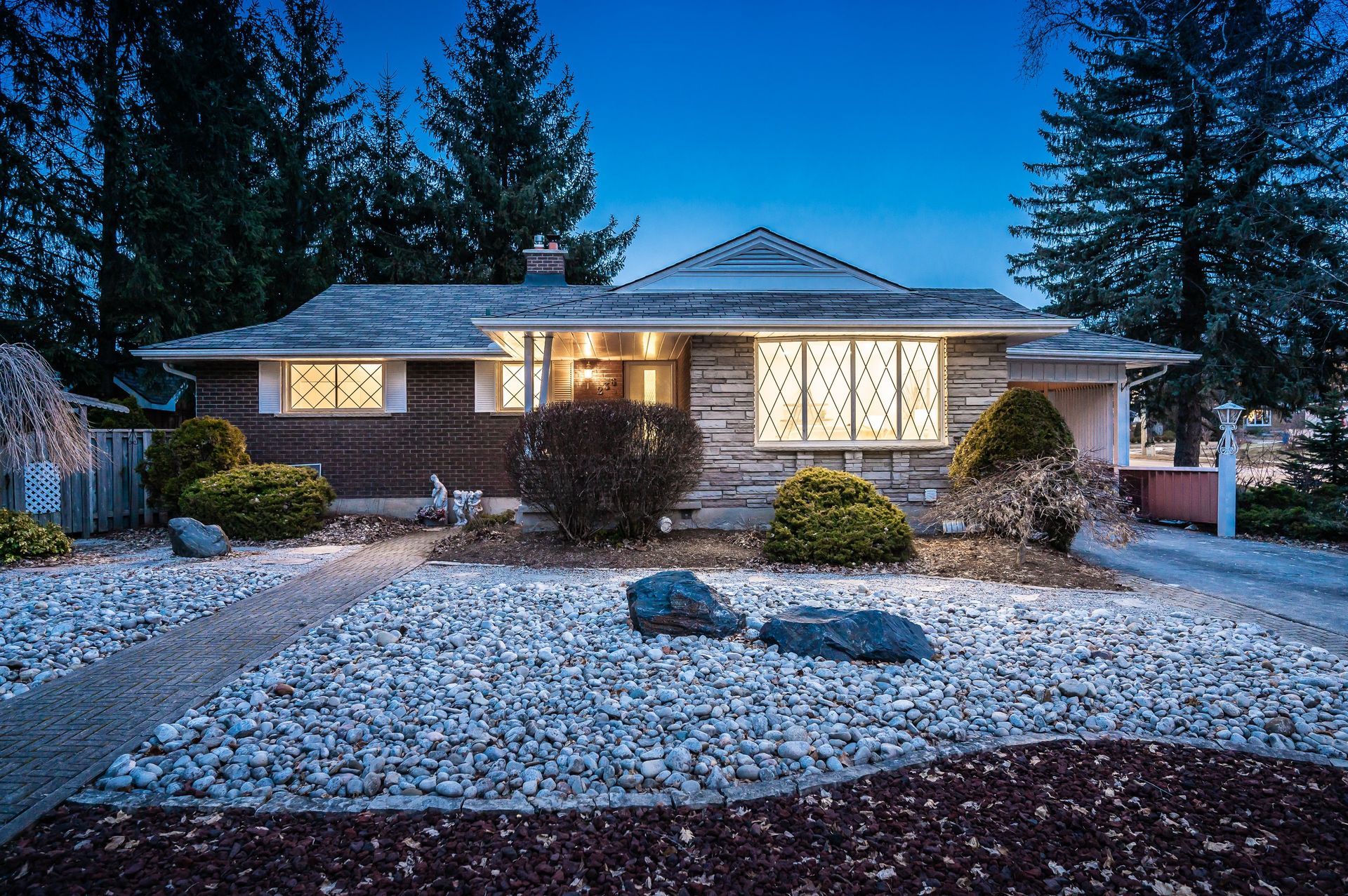 Ranch-style house at dusk with stone facade, manicured yard, and lit windows.