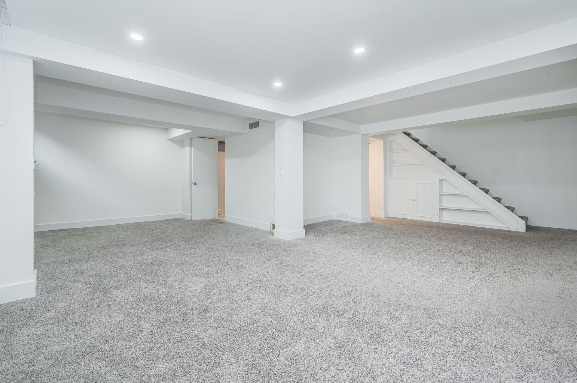 Empty, white-walled room with gray carpet, support beams, and recessed lighting. Stairs and doorway visible.