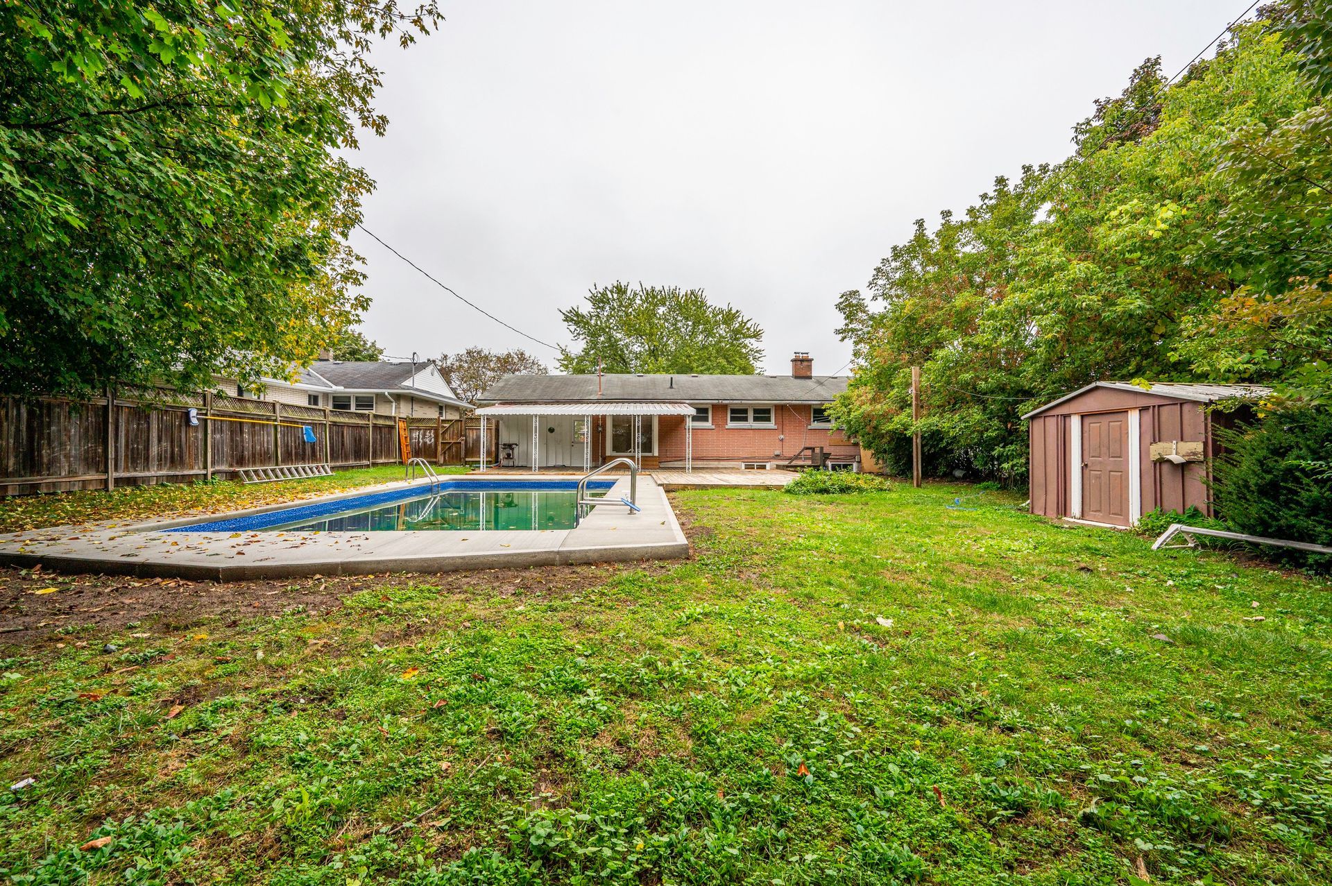 Backyard with pool, shed, green lawn, trees, and house. Overcast sky.