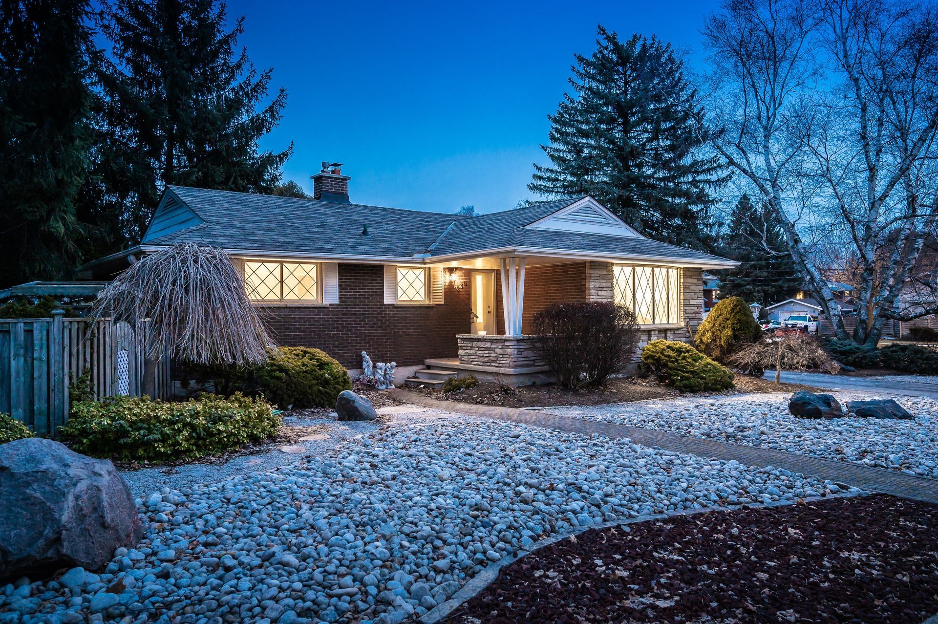 Ranch-style house at dusk, lit windows, stone pathway, landscaped yard with gravel and shrubs.