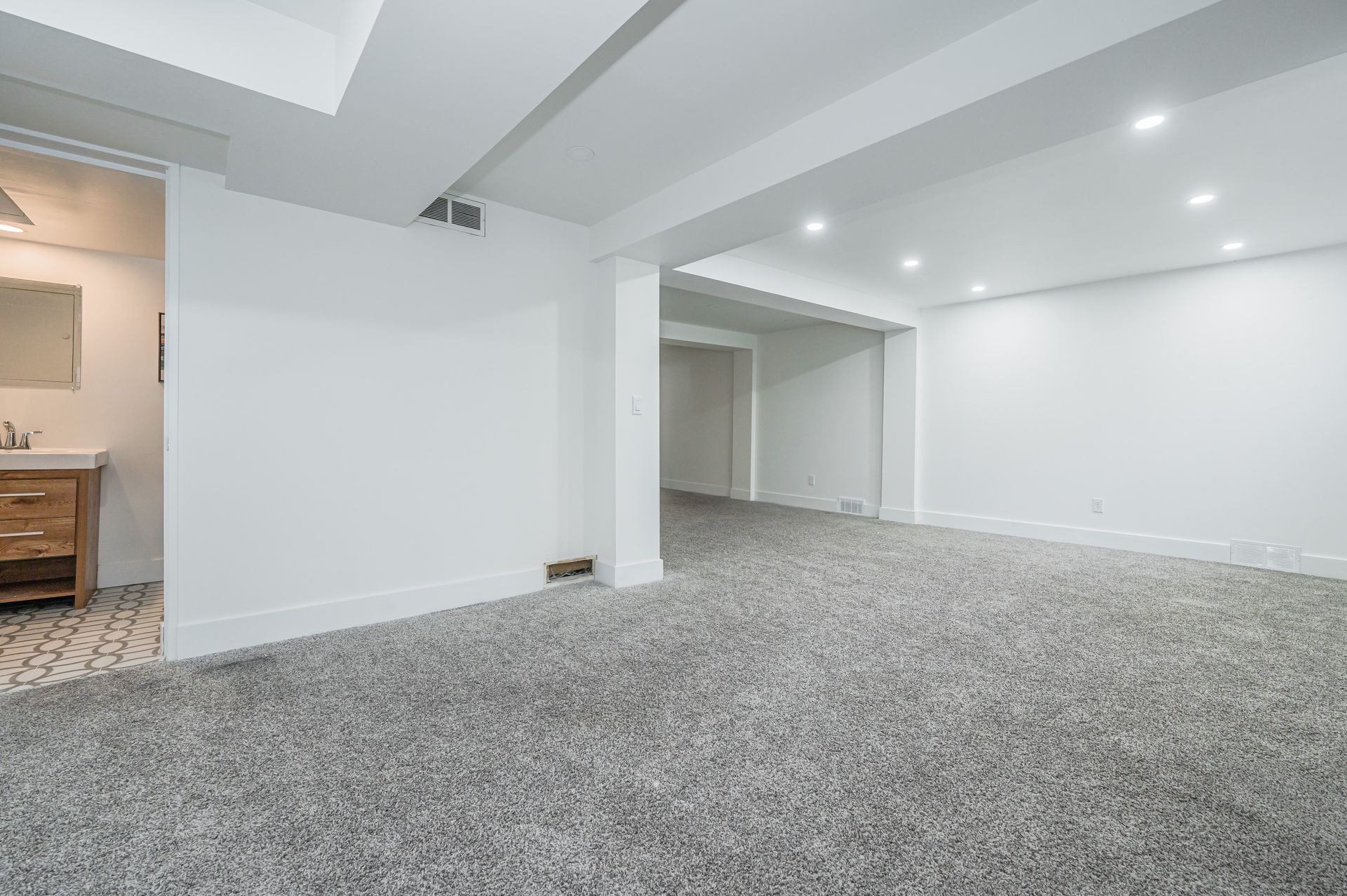 Empty white-walled basement with gray carpet. A bathroom doorway is visible on the left.