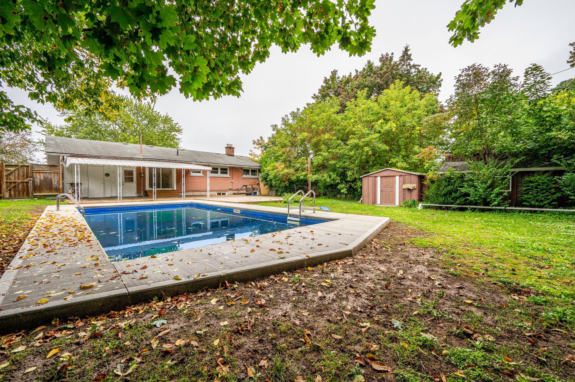Backyard with pool, shed, and green trees on a cloudy day.