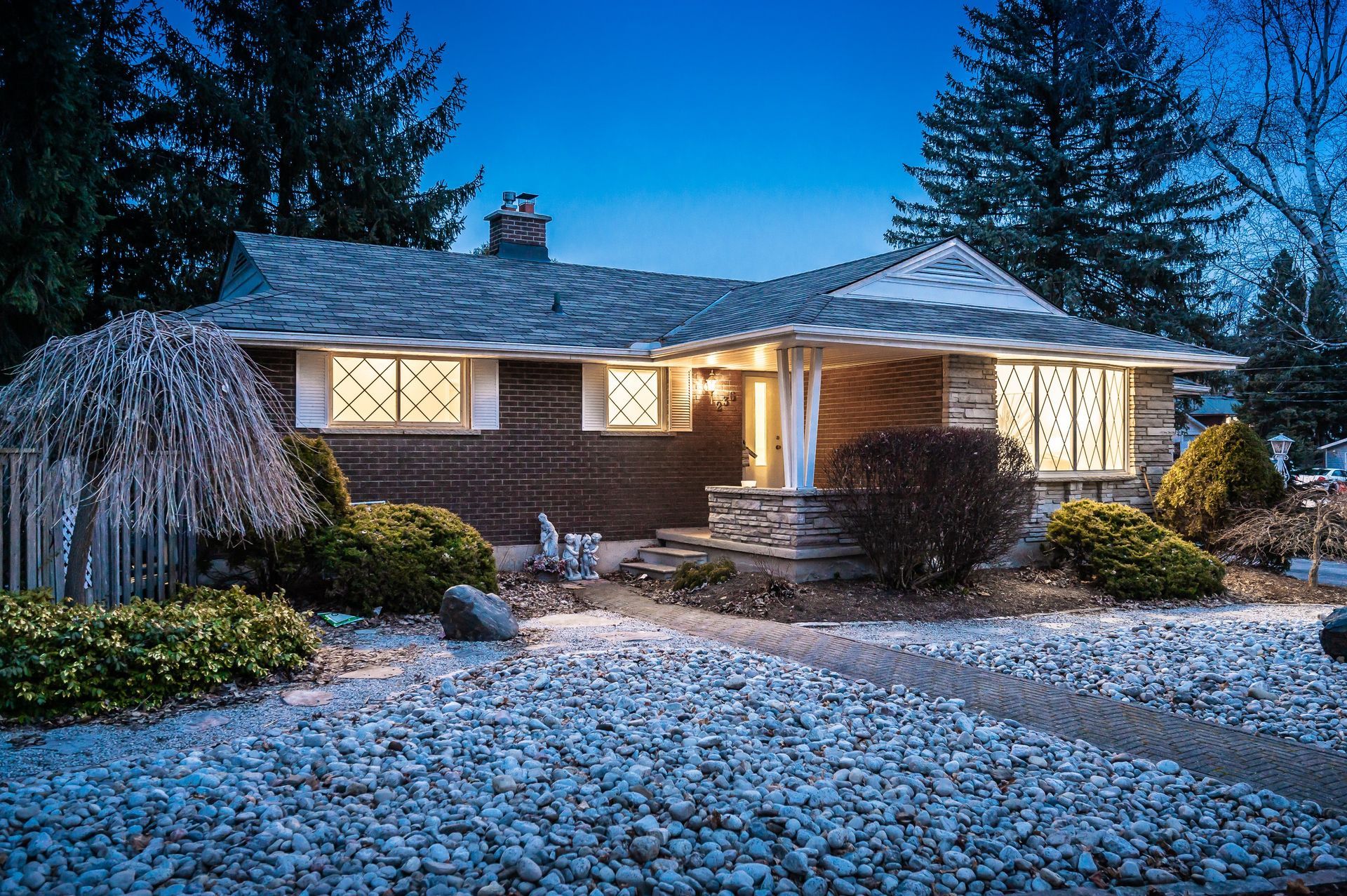 Ranch-style house at dusk with illuminated windows, stone facade, and landscaped front yard with gravel.