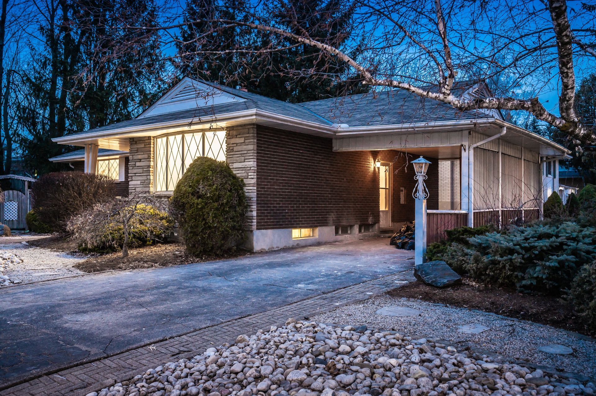 A single-story house at dusk, with a driveway, bushes, and a lantern.