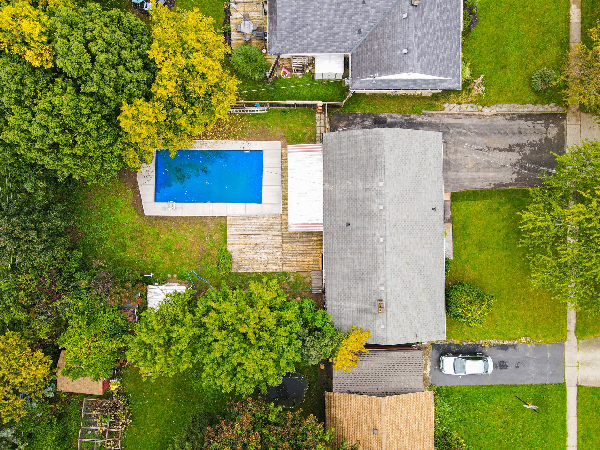 Overhead view of a house with a pool and a car parked in the driveway. Green lawn and trees surround the property.