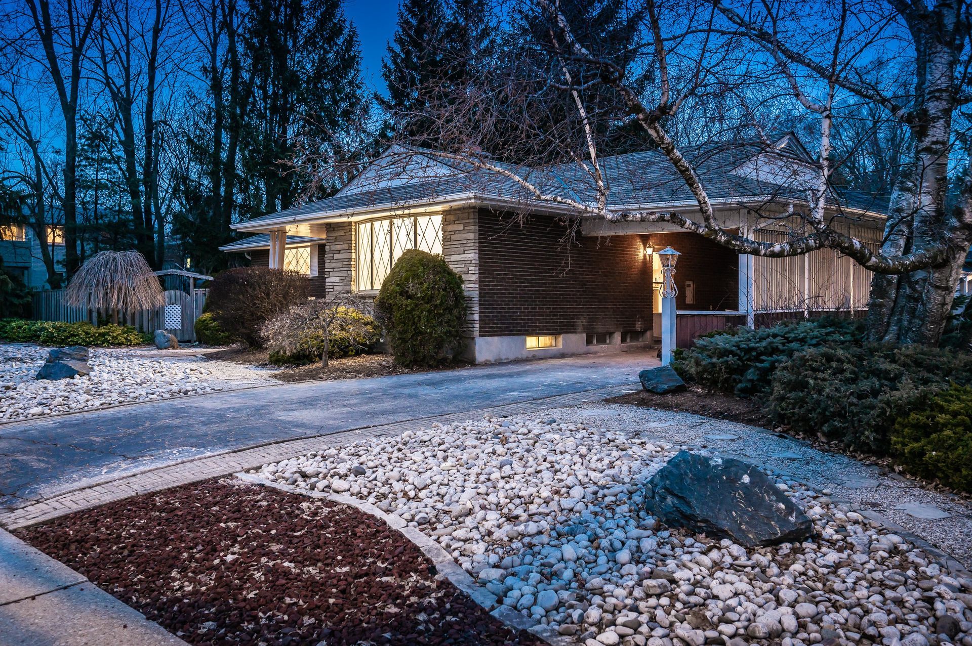 Snow-covered house at dusk, with illuminated windows and a carport. Driveway and landscaping visible.