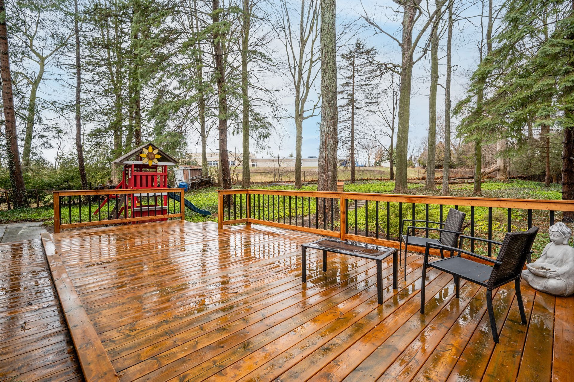 Wet wooden deck with black chairs and table, overlooking a backyard with trees and a small playhouse.