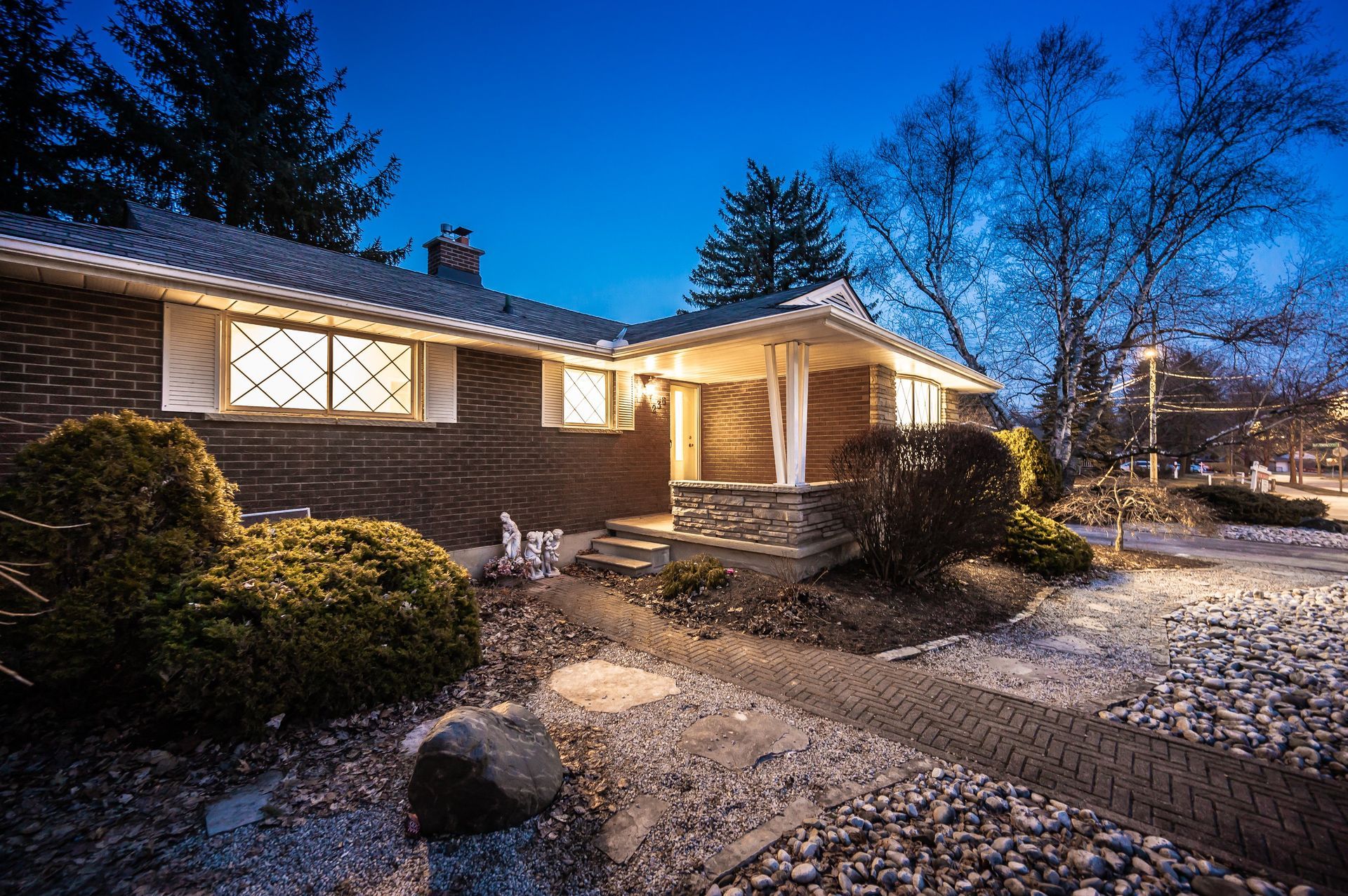 Evening view of a brick house with lights on, a stone walkway, and landscaping.