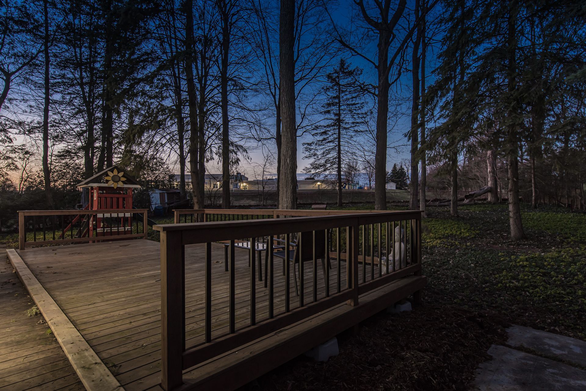Wooden deck in a wooded area at dusk with a children's playhouse in the distance.