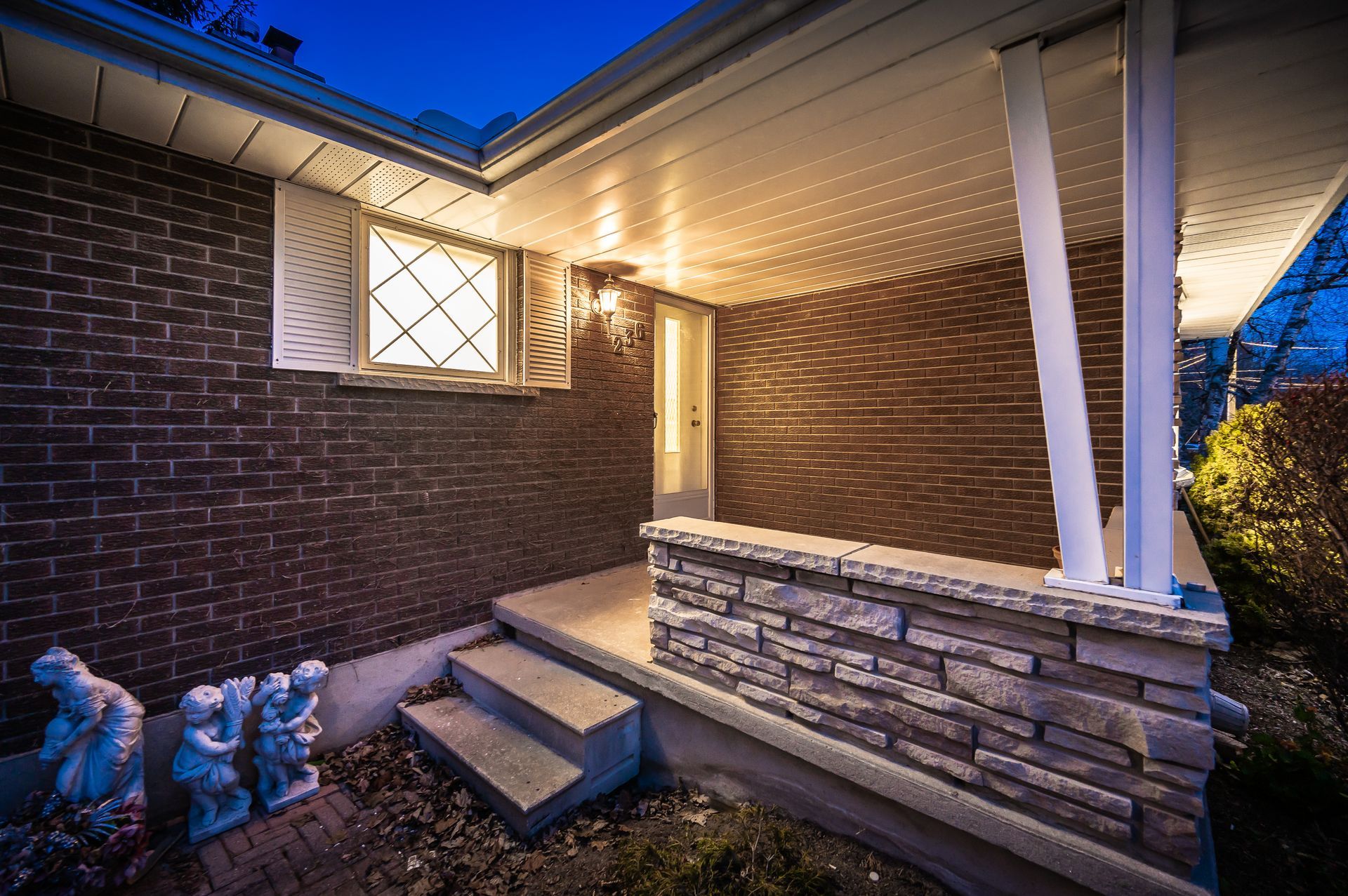Brick house entrance with stone-faced wall, concrete steps, white porch columns, and a lit doorway at dusk.