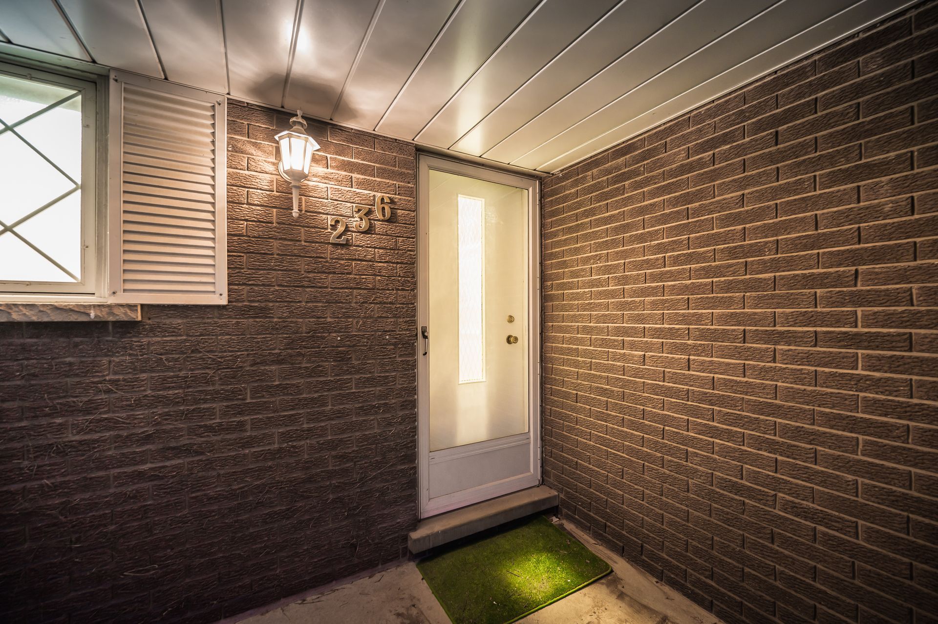 Brick-walled entryway with a white door, window with shutters, and a green welcome mat.