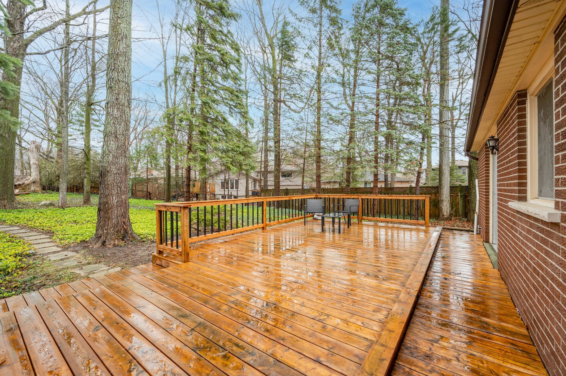 Wooden deck with black railing next to a brick building, surrounded by tall trees. Wet from rain.