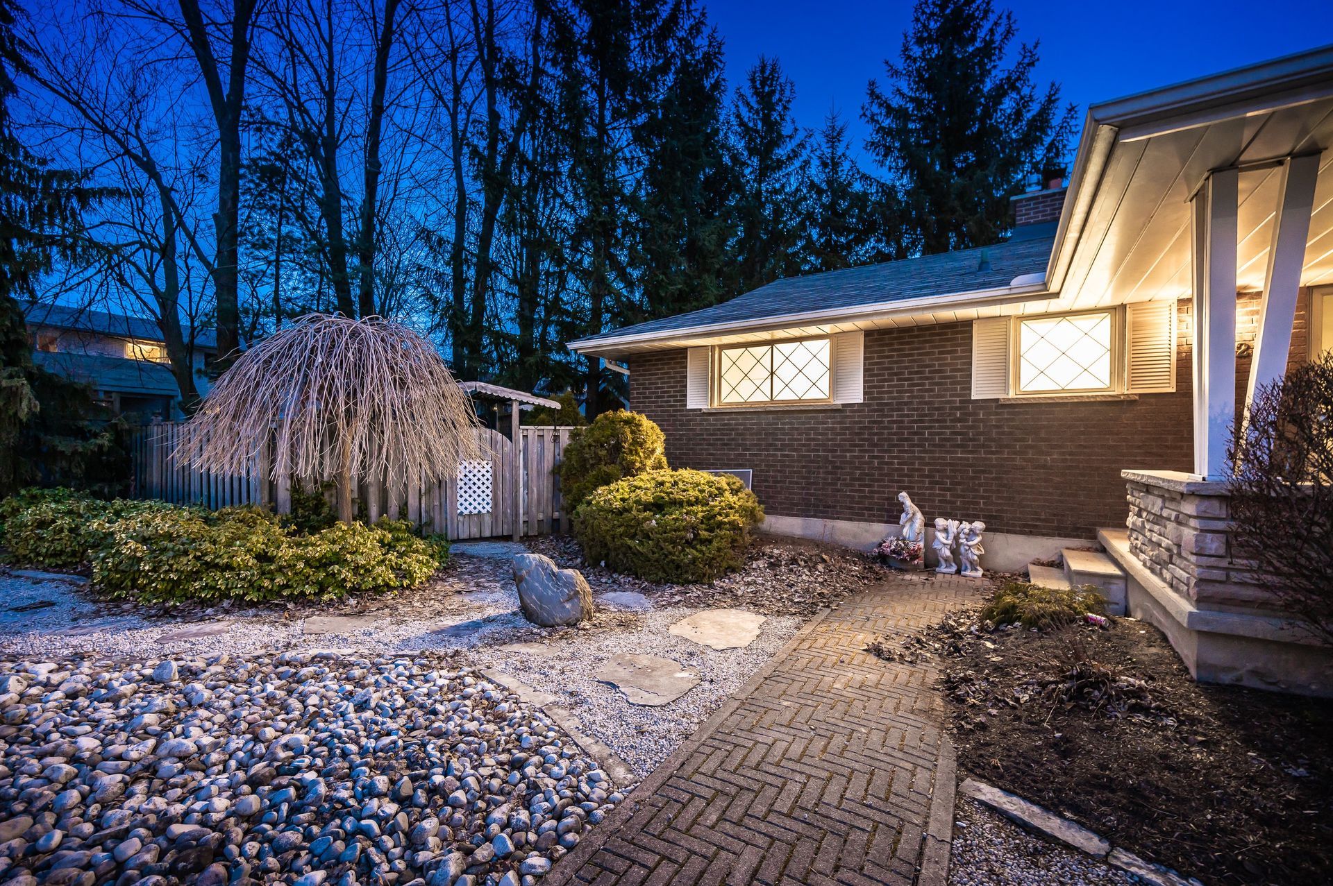 Night exterior of house with pathway, landscaping, and lit porch. Dark brick, trees.