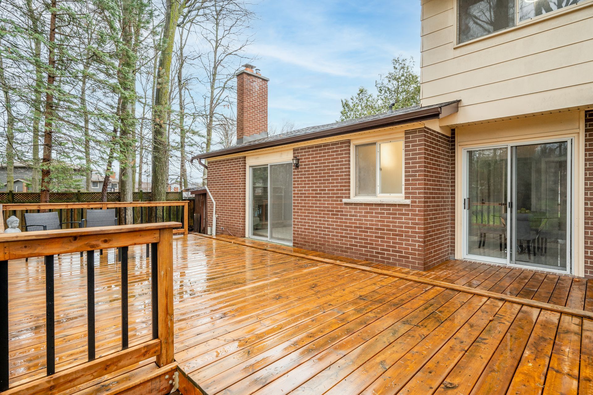 Wooden deck attached to a brick house with sliding glass doors.  Green trees in the background, cloudy sky.
