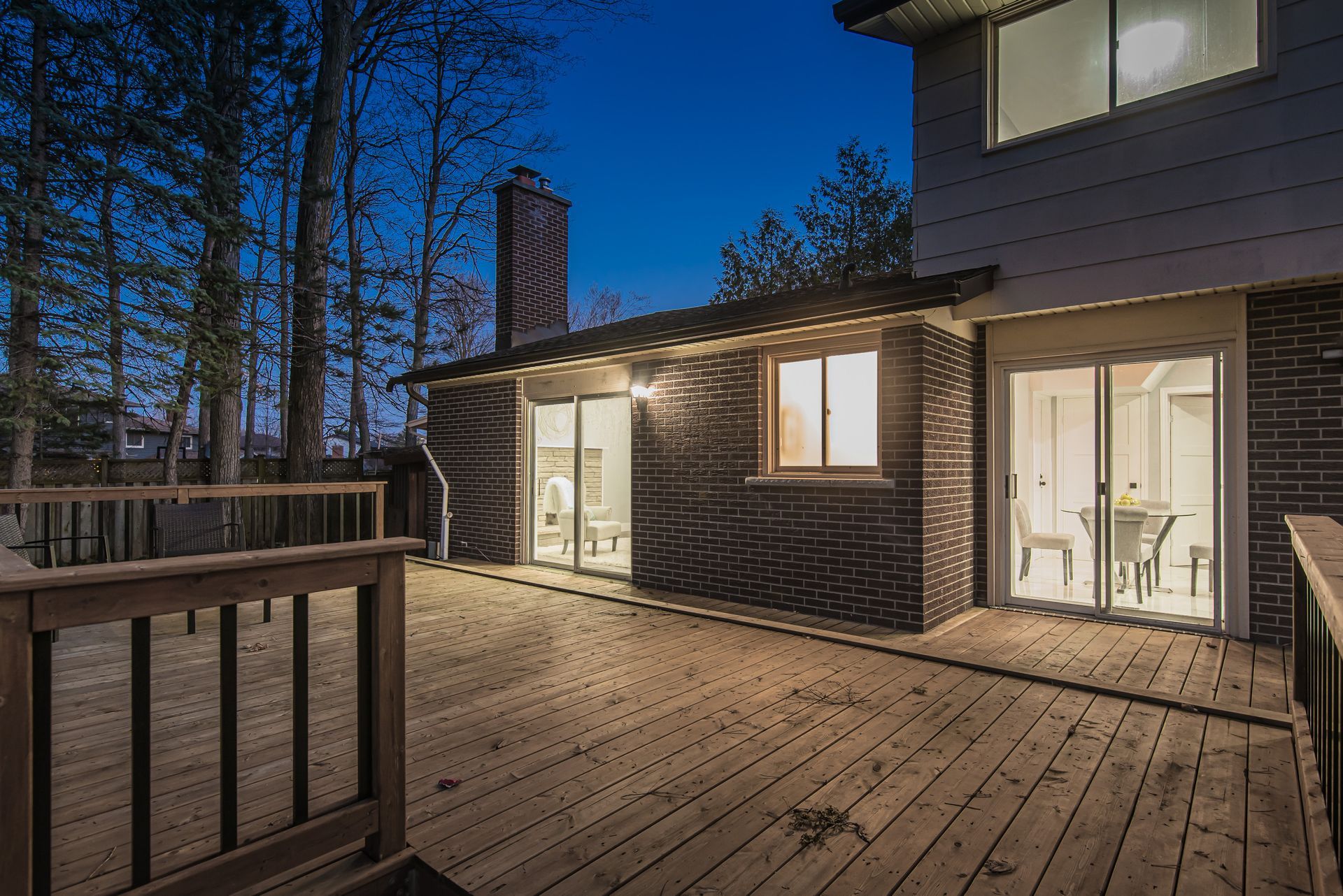 Wooden deck at dusk; brick house with lit windows and chimney; trees in background.