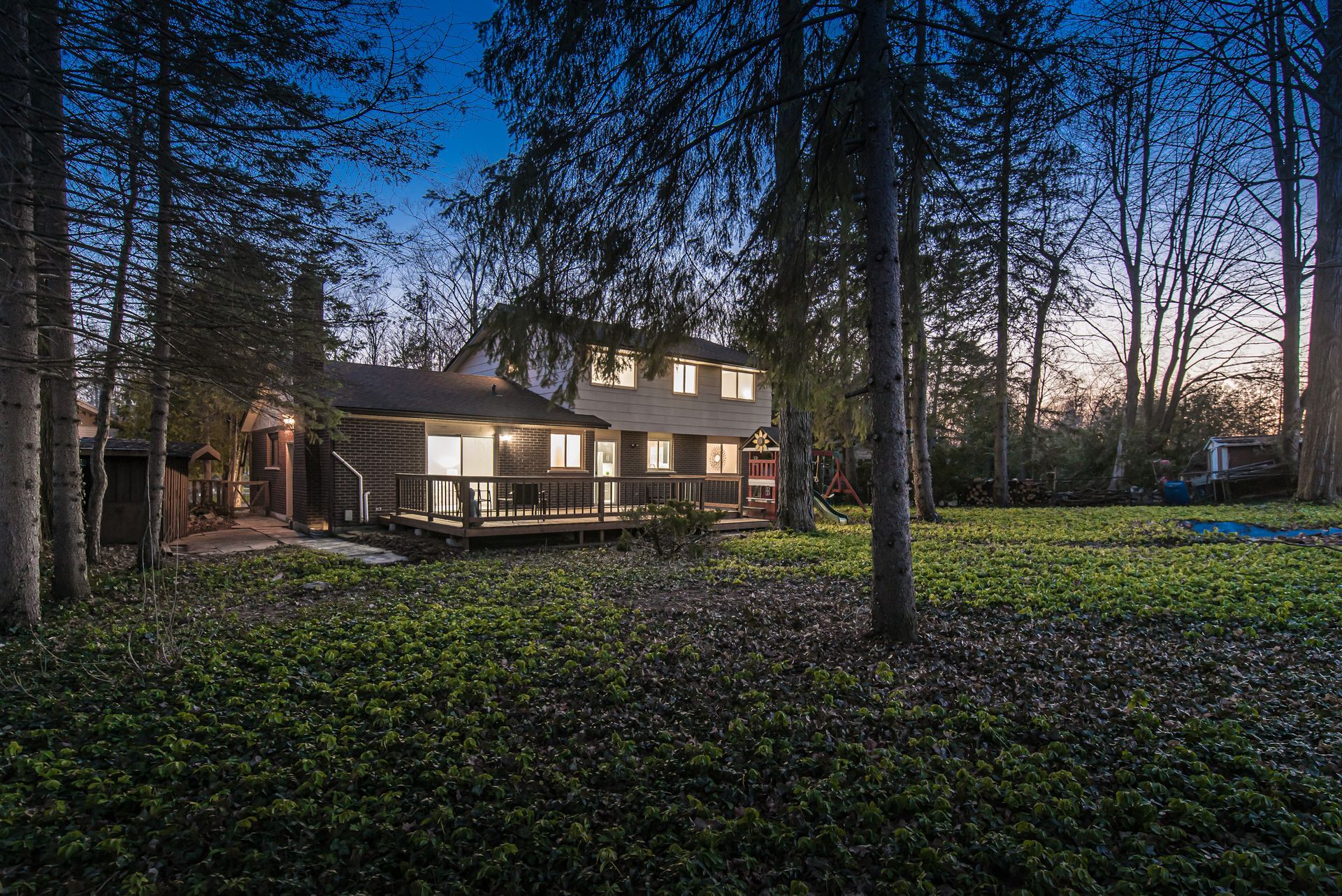 House exterior at dusk, with lit windows and deck, surrounded by trees and a grassy yard.