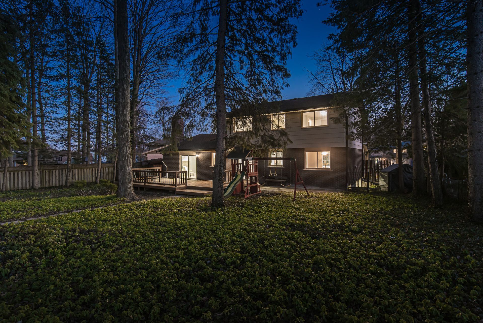 Backyard view of a two-story house at dusk, with trees, a deck, and yard lights.