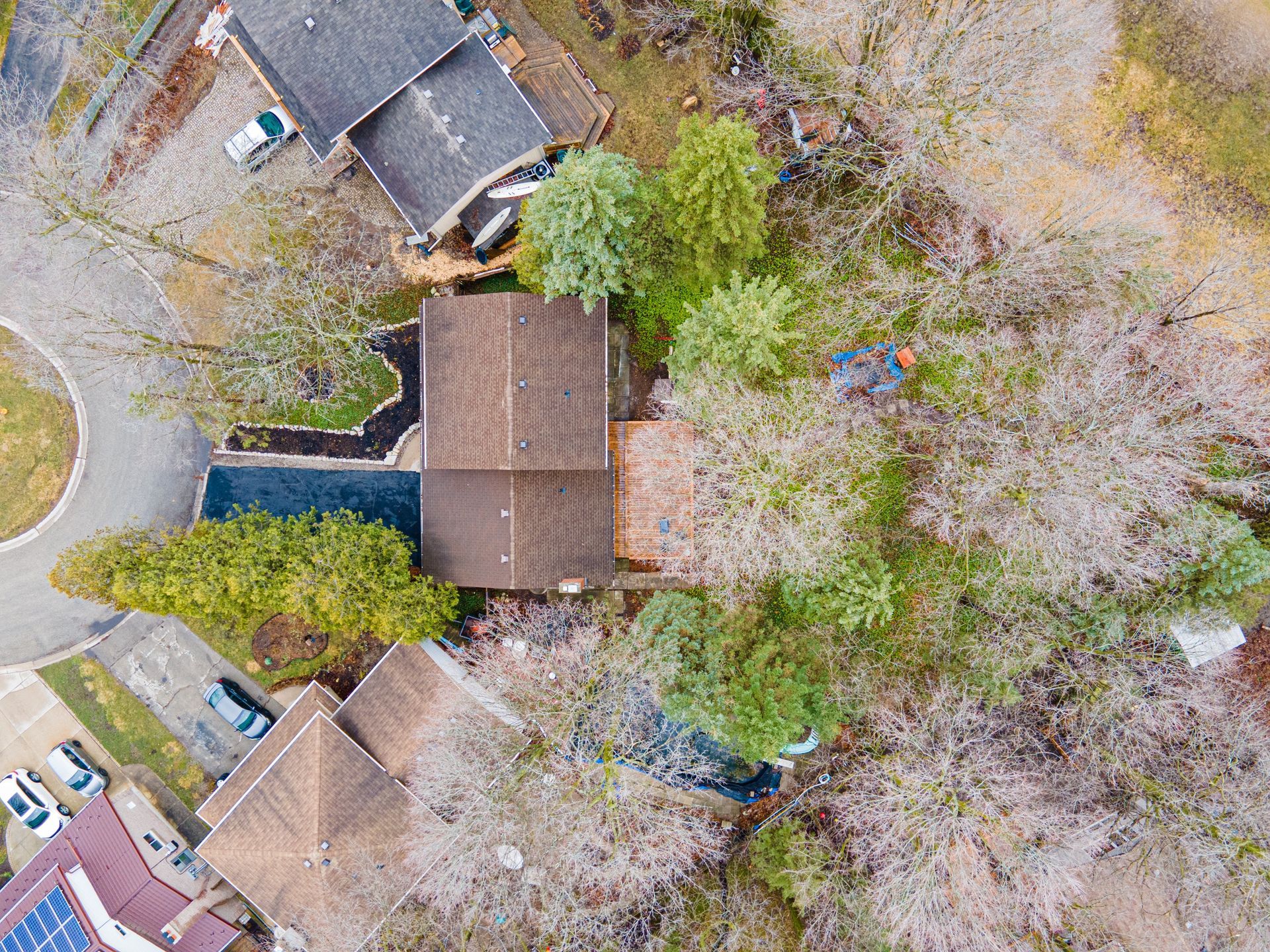 Overhead view of several houses with brown roofs surrounded by trees and some cars parked on driveways.